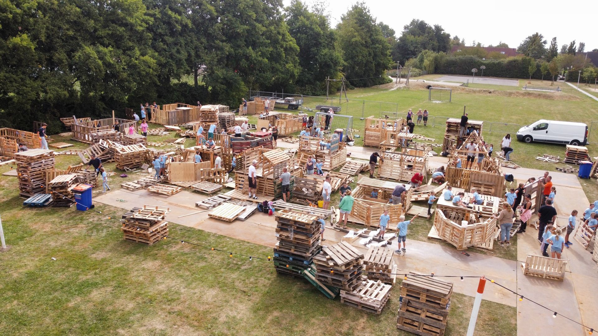 Mensen bouwen constructies van houten pallets op een grasveld; bomen op de achtergrond.