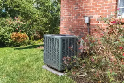 Outdoor air conditioner beside a brick house, surrounded by green lawn and shrubs.