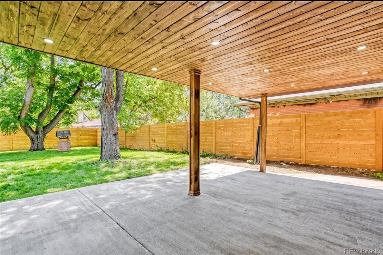 Covered concrete patio with wooden ceiling, central post, and fenced yard with trees and grass