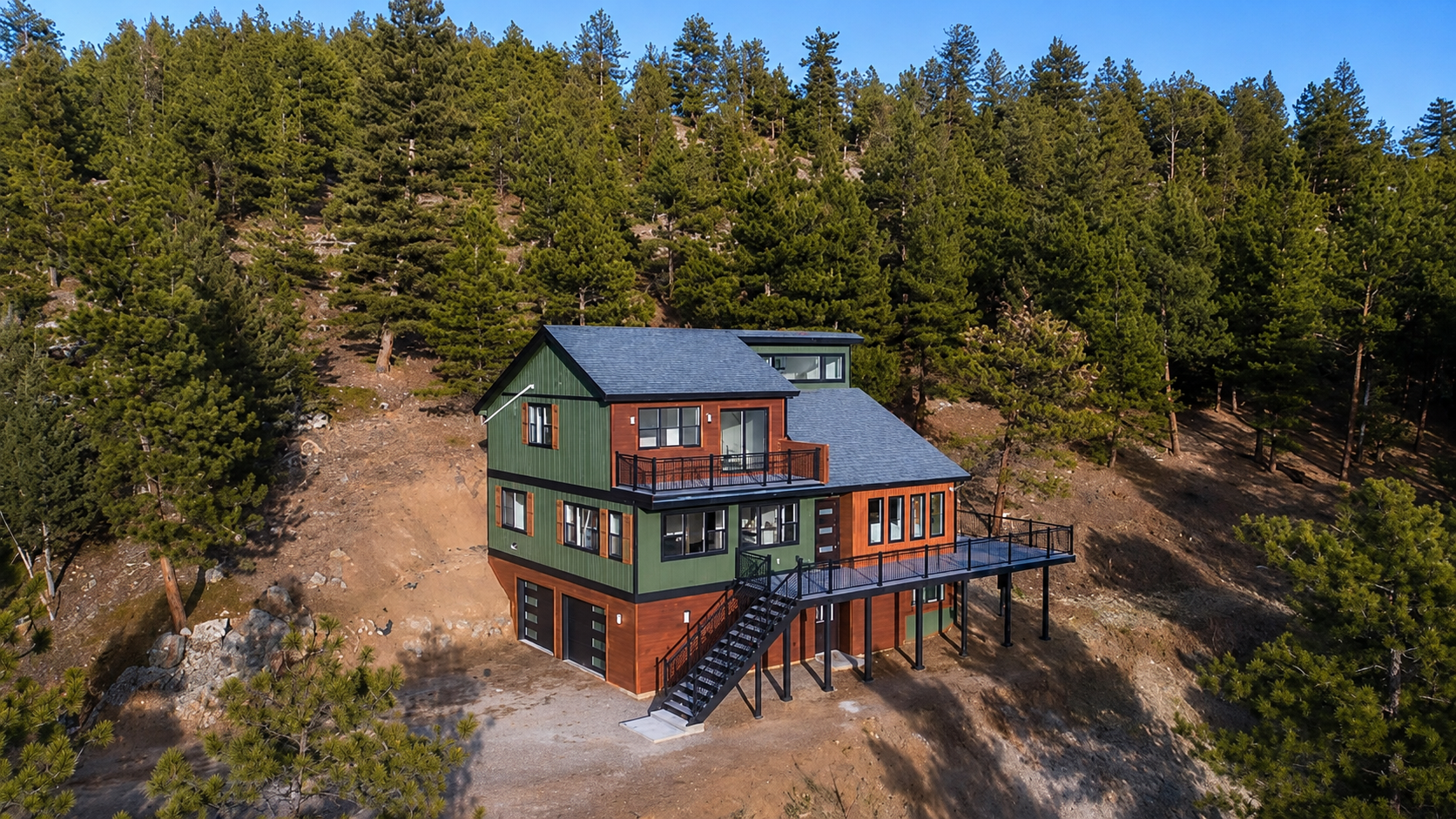 Modern two-story cabin with green roof and wood deck on a hillside surrounded by pine forest