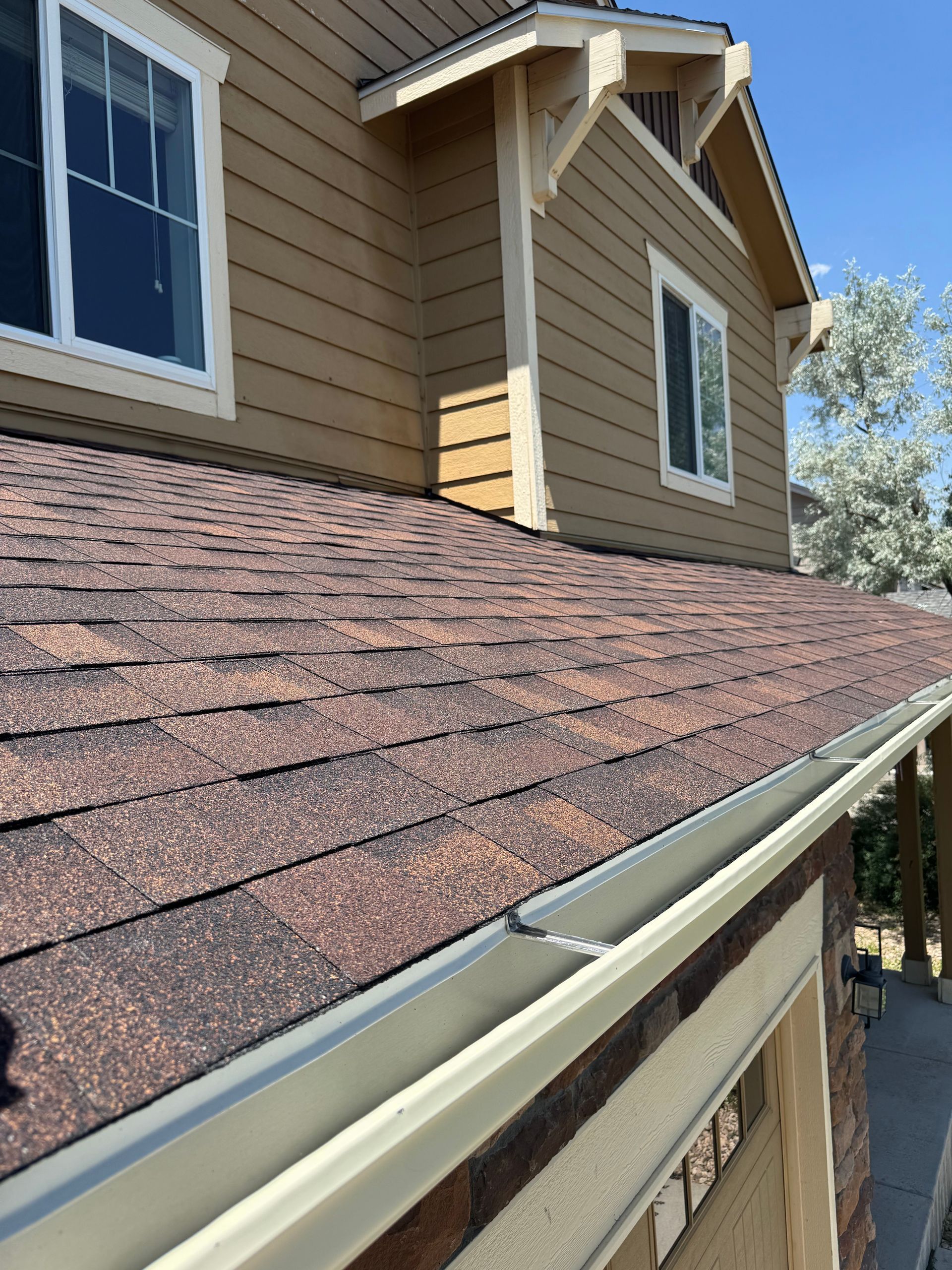 House exterior with brown shingle roof and beige siding, seen from a rain gutter and fence below