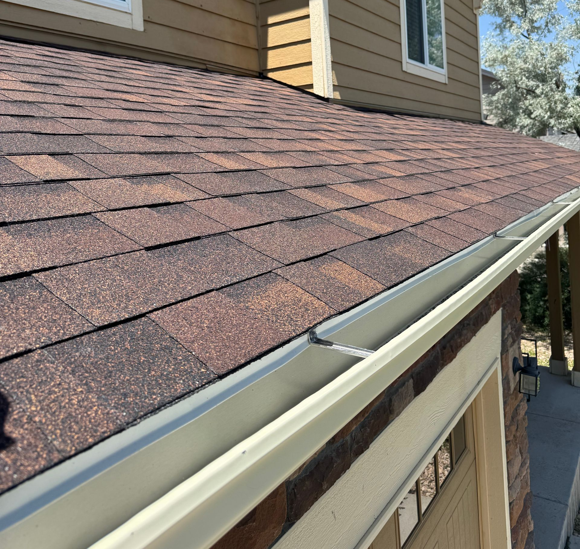 Brown shingle roof with white gutter and tan house siding on a sunny day