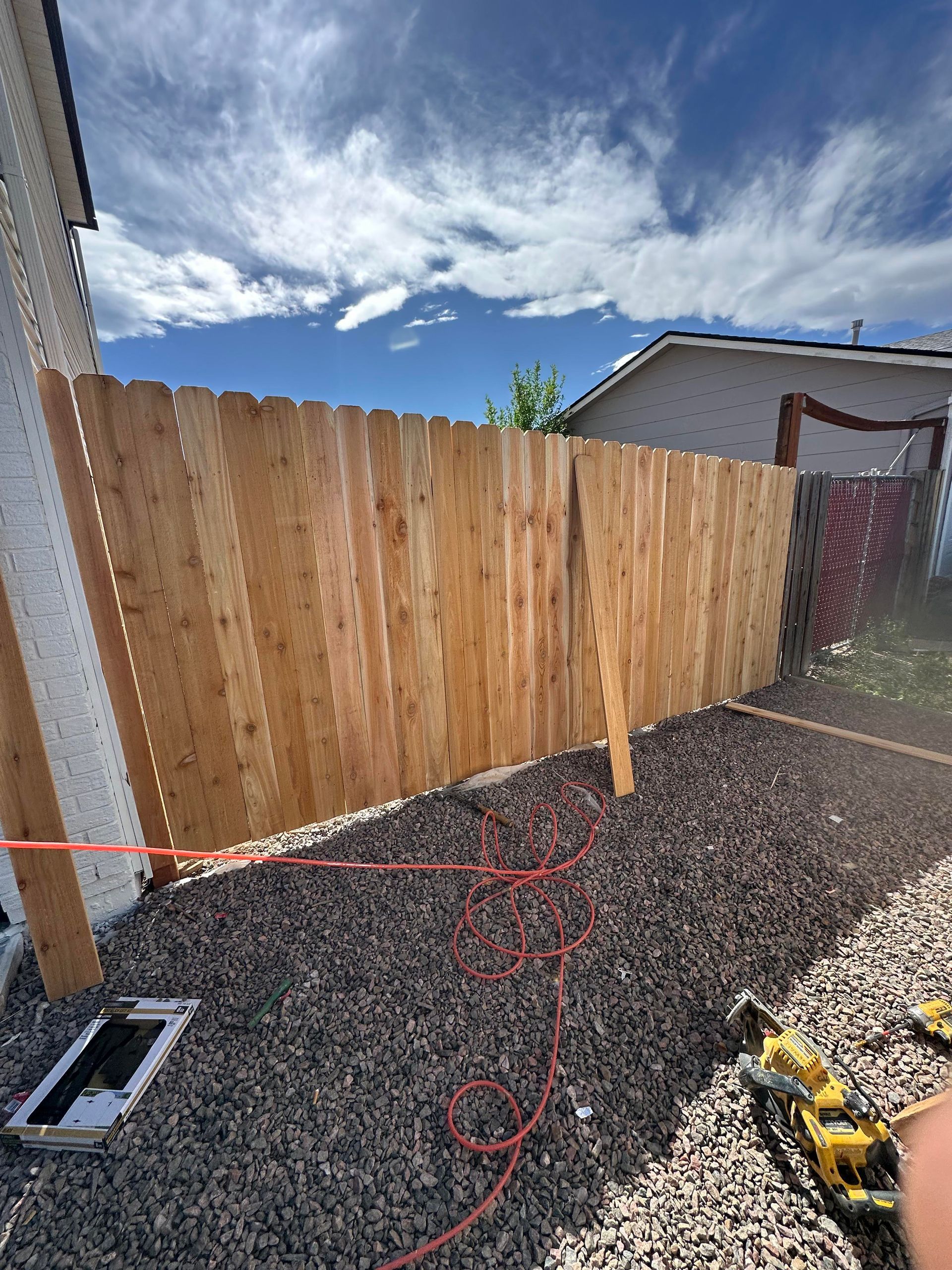 Backyard with new wooden fence, gravel ground, blue sky, and a few tools and cables in the foreground