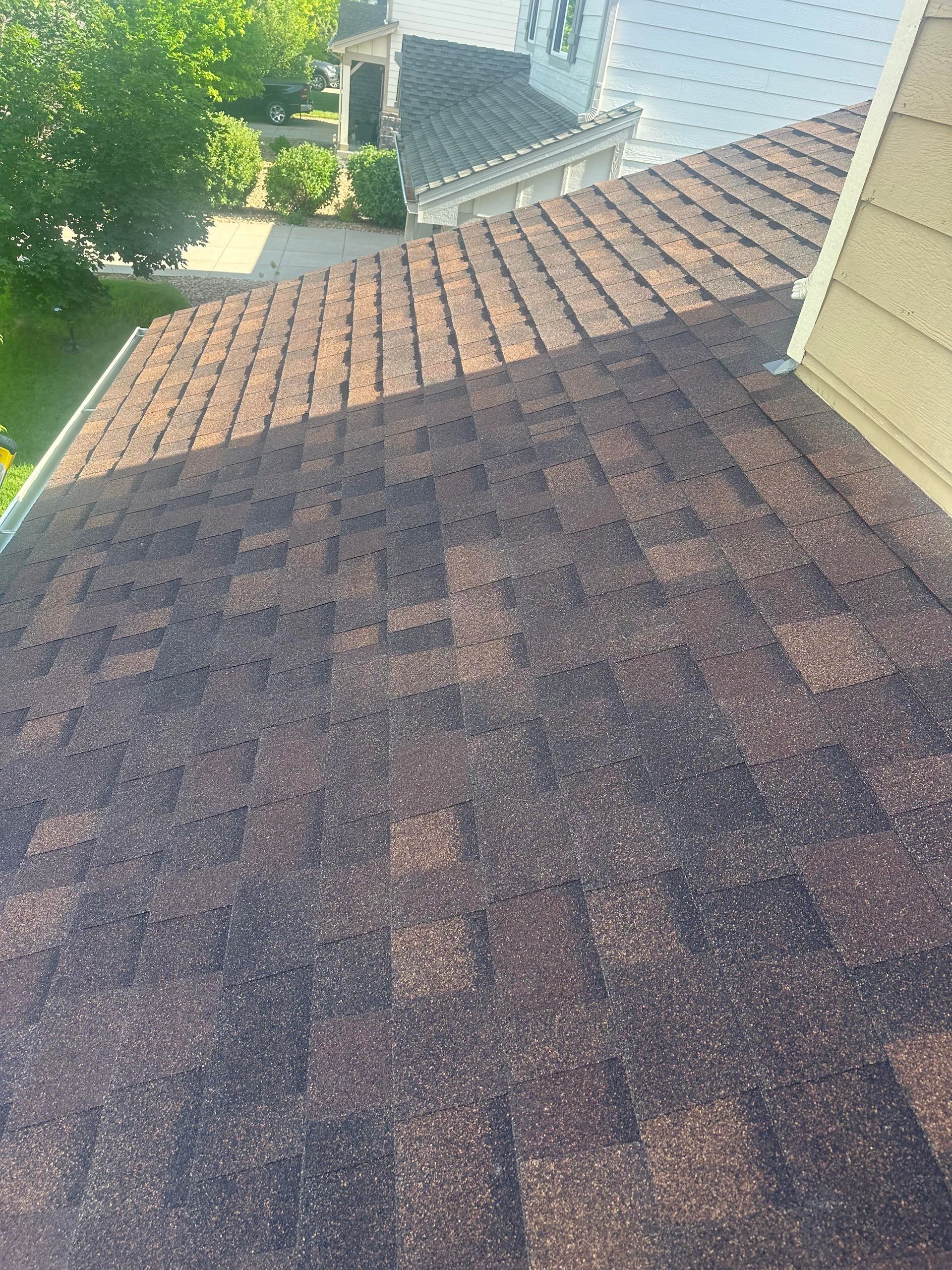 Shingled roof with brown and gray tiles in sunlight, beside a house and leafy trees