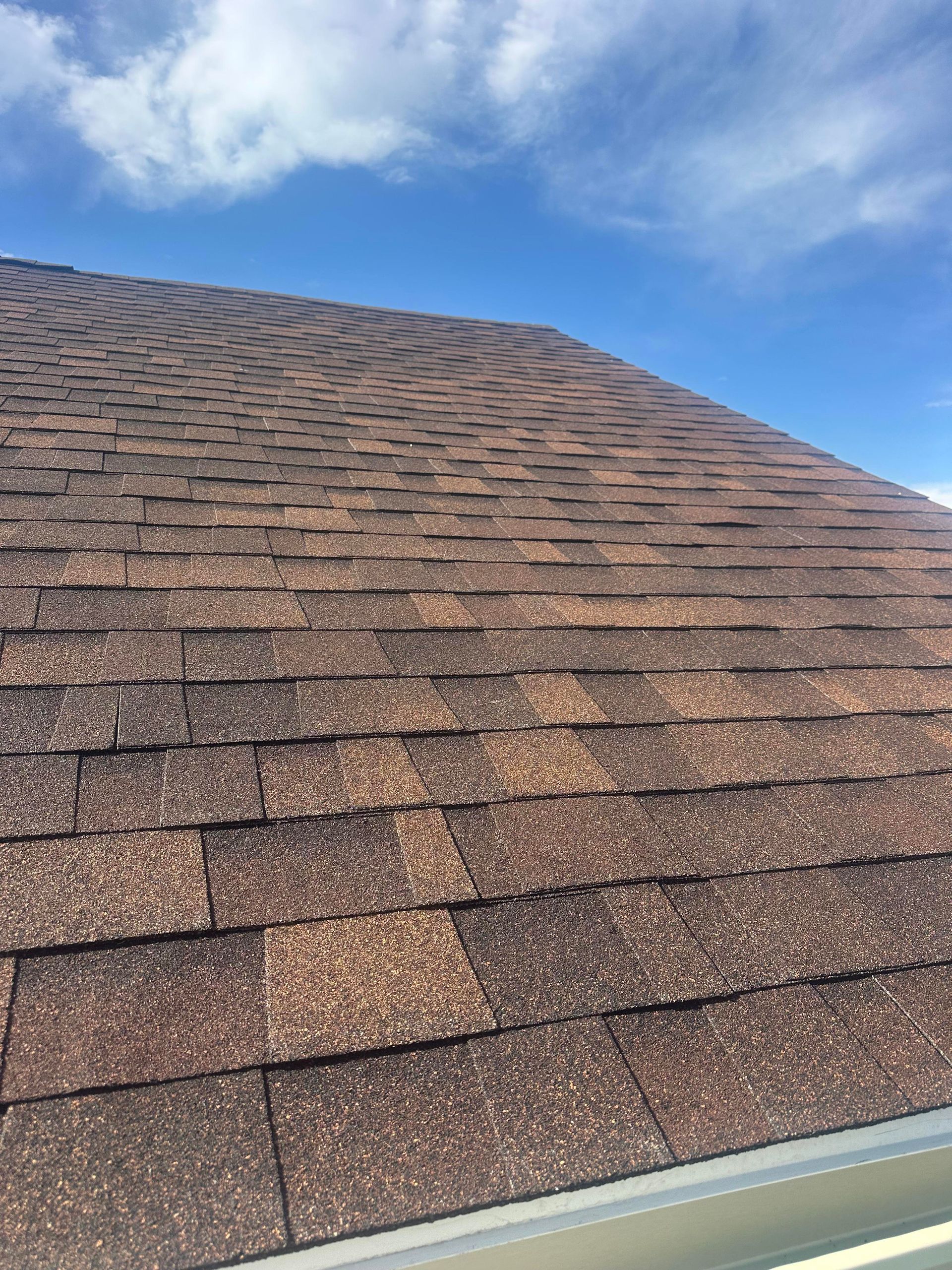 Brown shingled roof with a white gutter against a blue sky with clouds