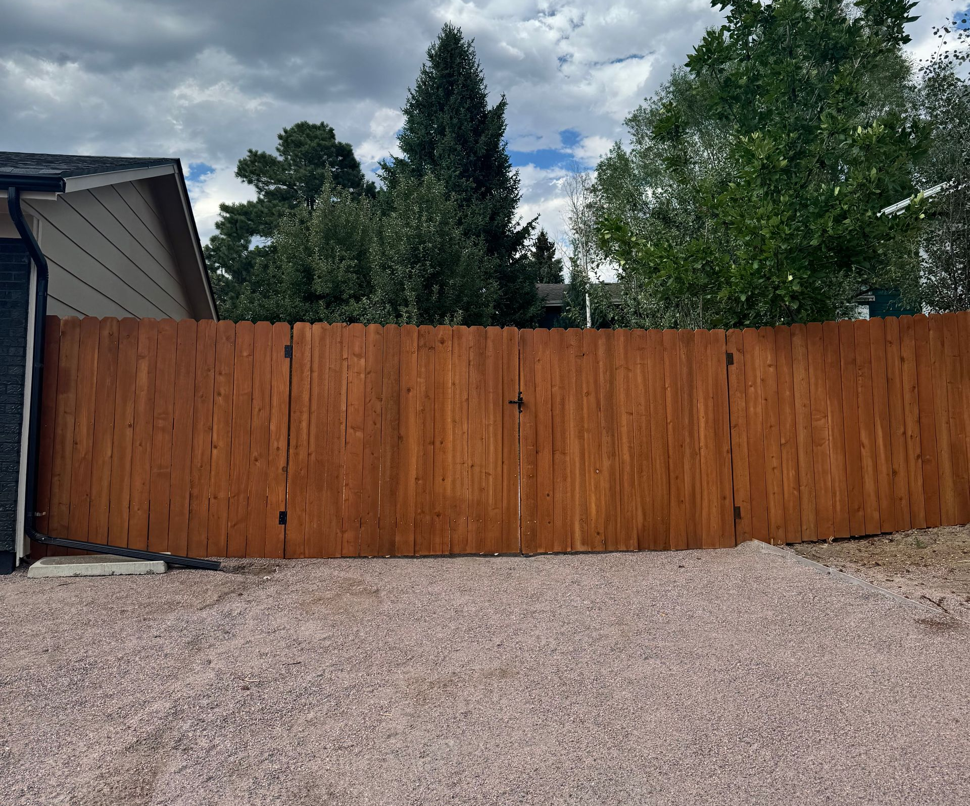 Wooden privacy fence beside a gravel driveway under cloudy skies, with trees and part of a house visible.