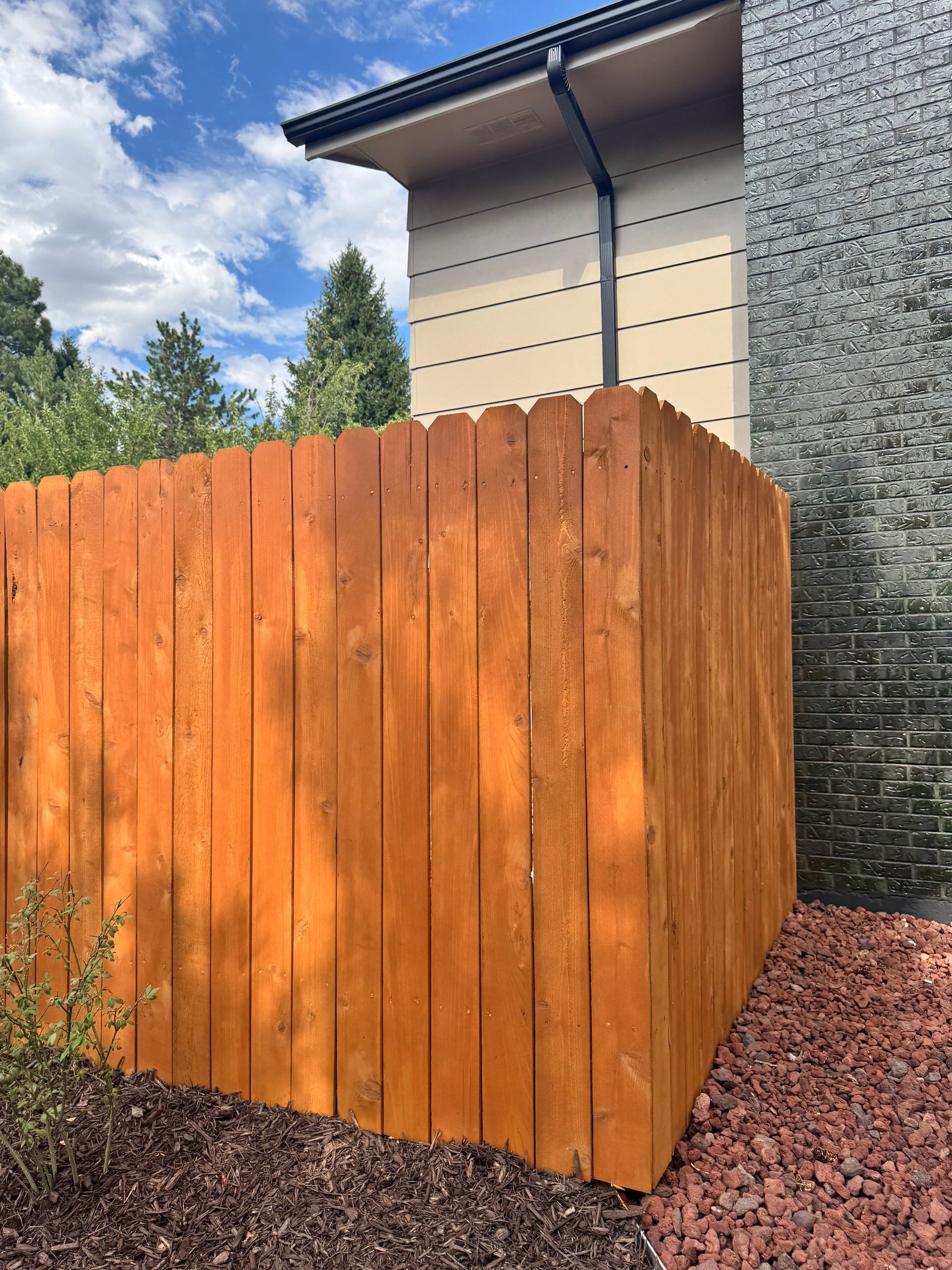 Wooden slatted fence beside a house, with gravel and trees under a blue sky.