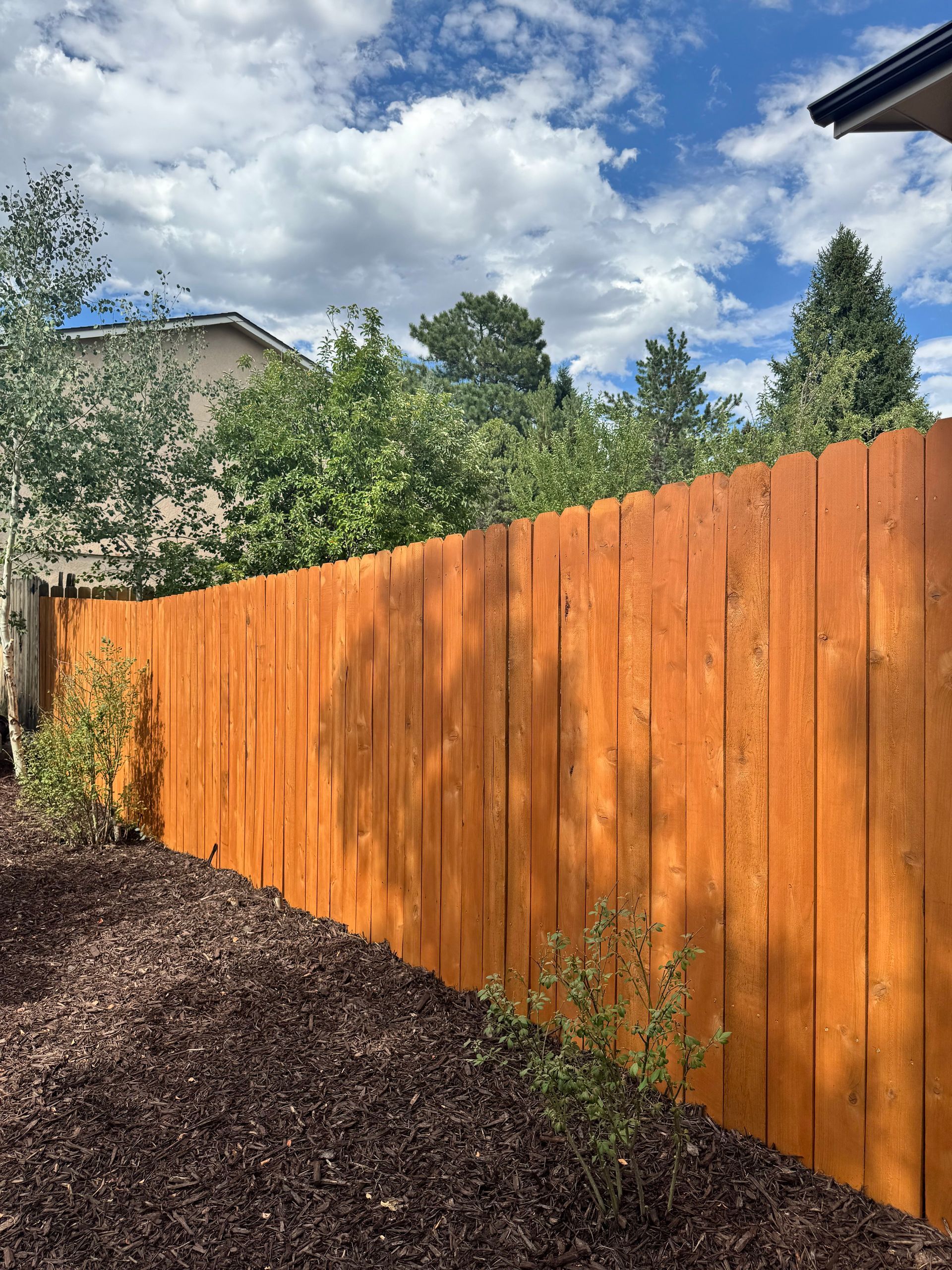 Orange wooden fence along a mulch-covered yard under a cloudy sky with trees and a house in the background