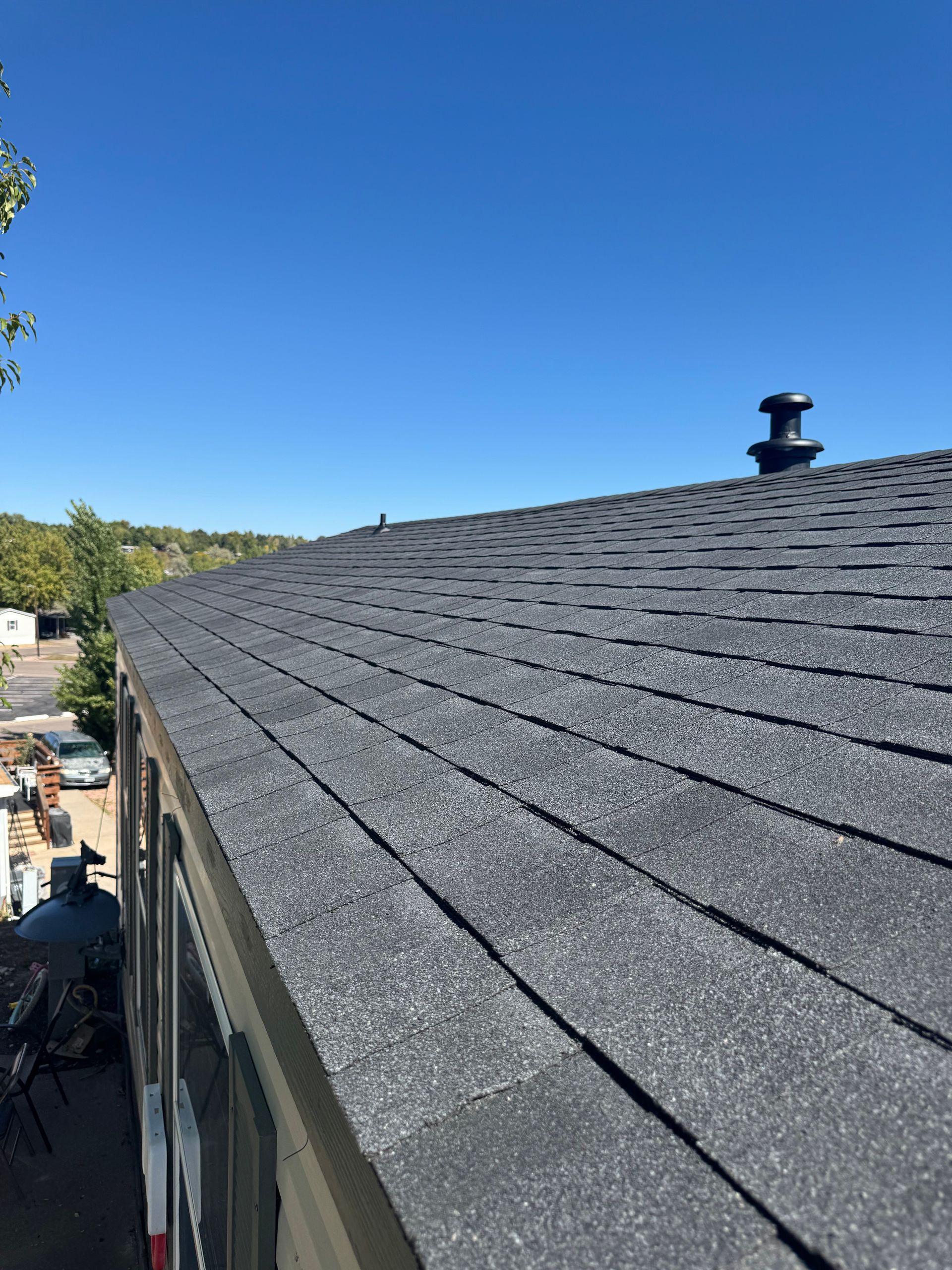 Shingled roof of a house under a clear blue sky, with a small vent pipe near the ridge.