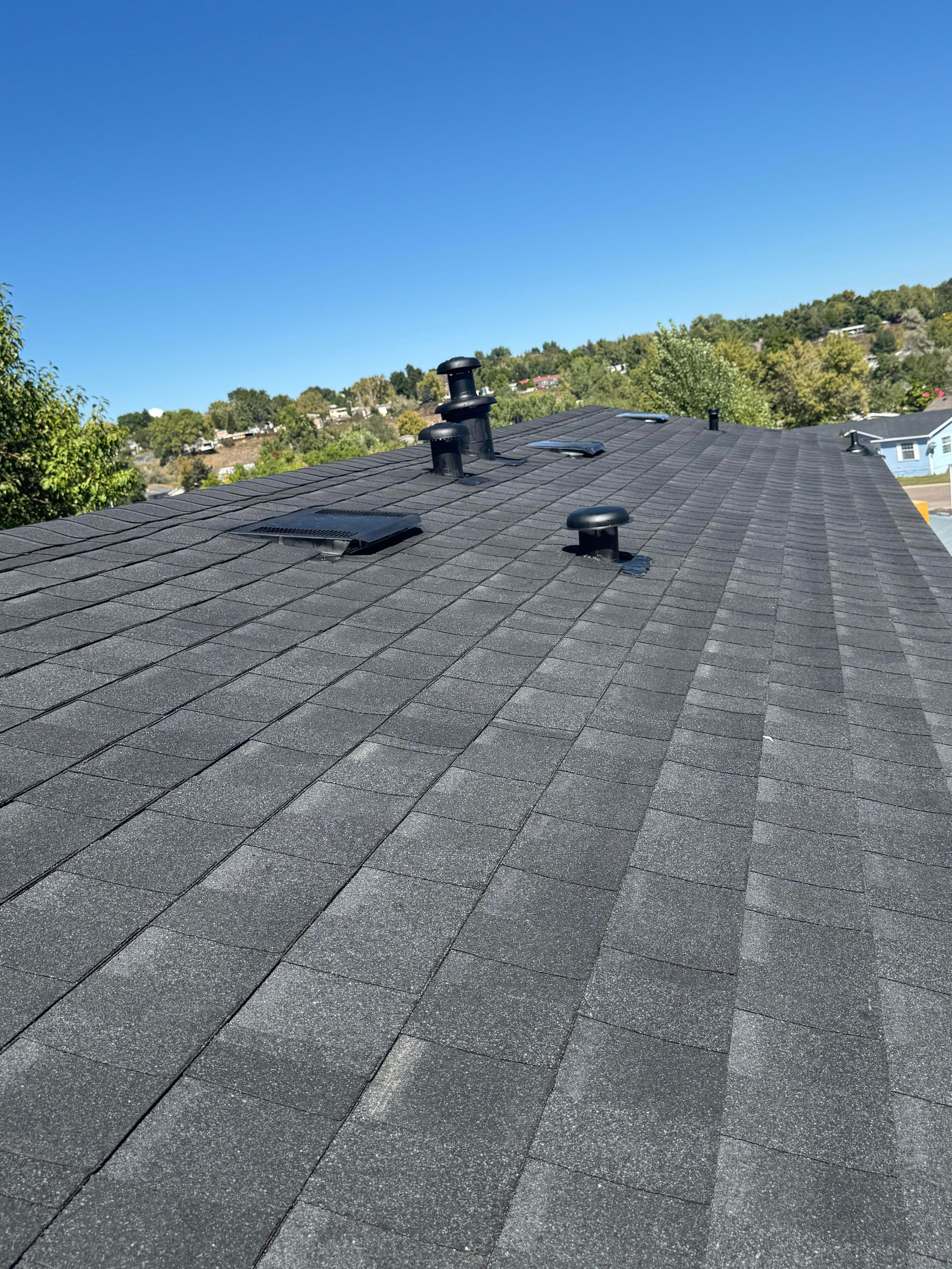 Rooftop with dark shingles, two vents, and a clear blue sky over a hilly neighborhood