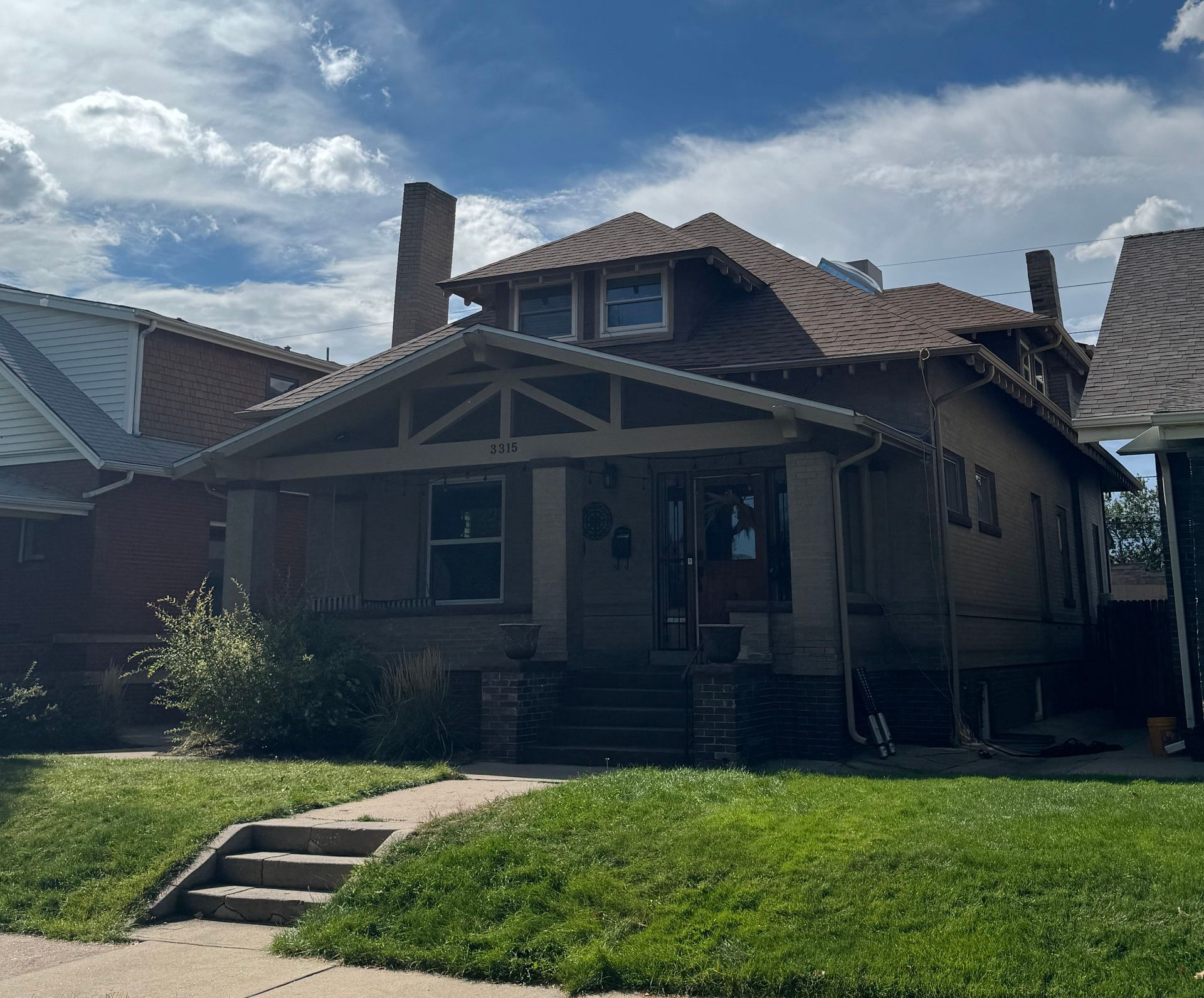 Front view of a small beige bungalow with a front porch, chimney, and green lawn under a partly cloudy sky