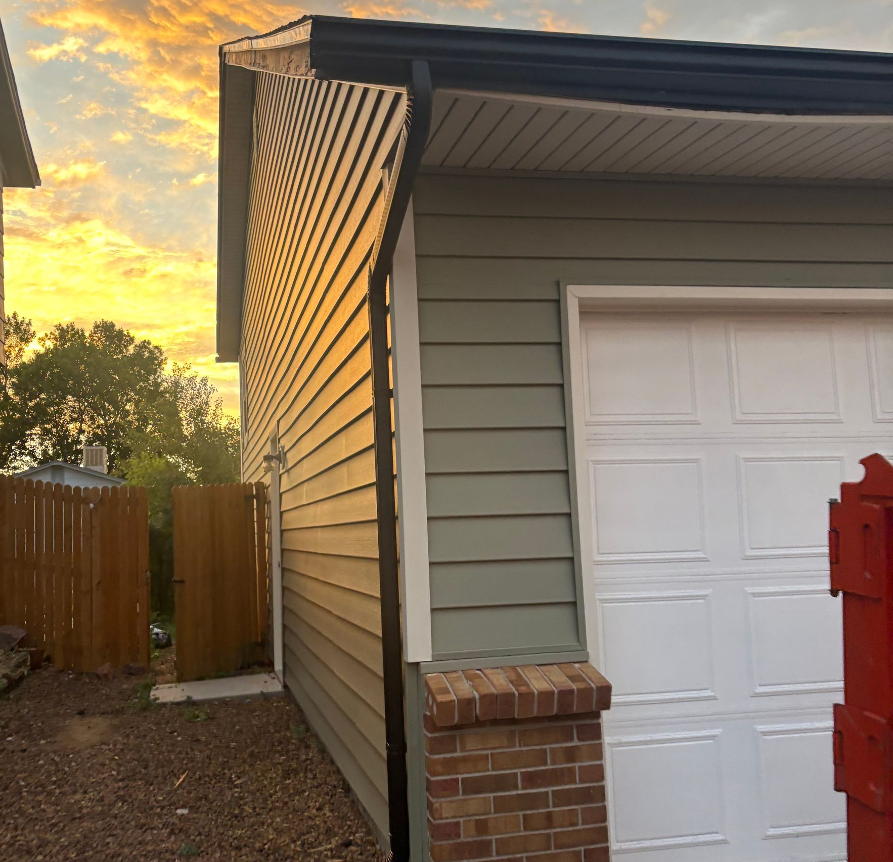 Sunset view of a house side wall, garage door, wooden fence, and red mailbox near a brick corner.