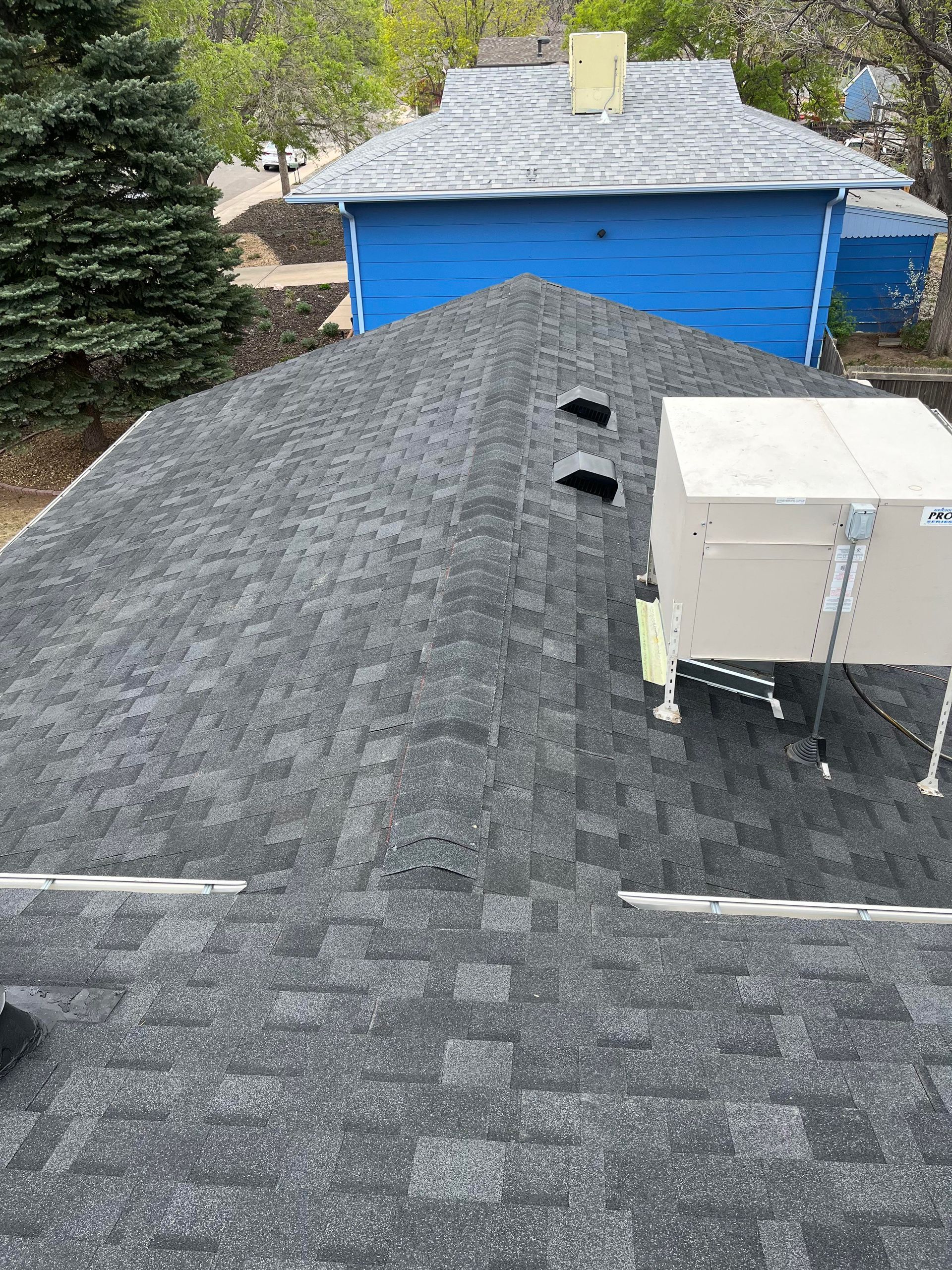 Rooftop with dark shingles, two vents, and a white HVAC unit, with a blue house and tree in the background