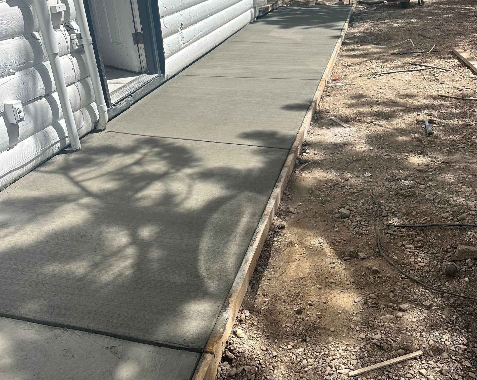 Concrete sidewalk beside a white building, with dirt and gravel along the edge and shadows across the path