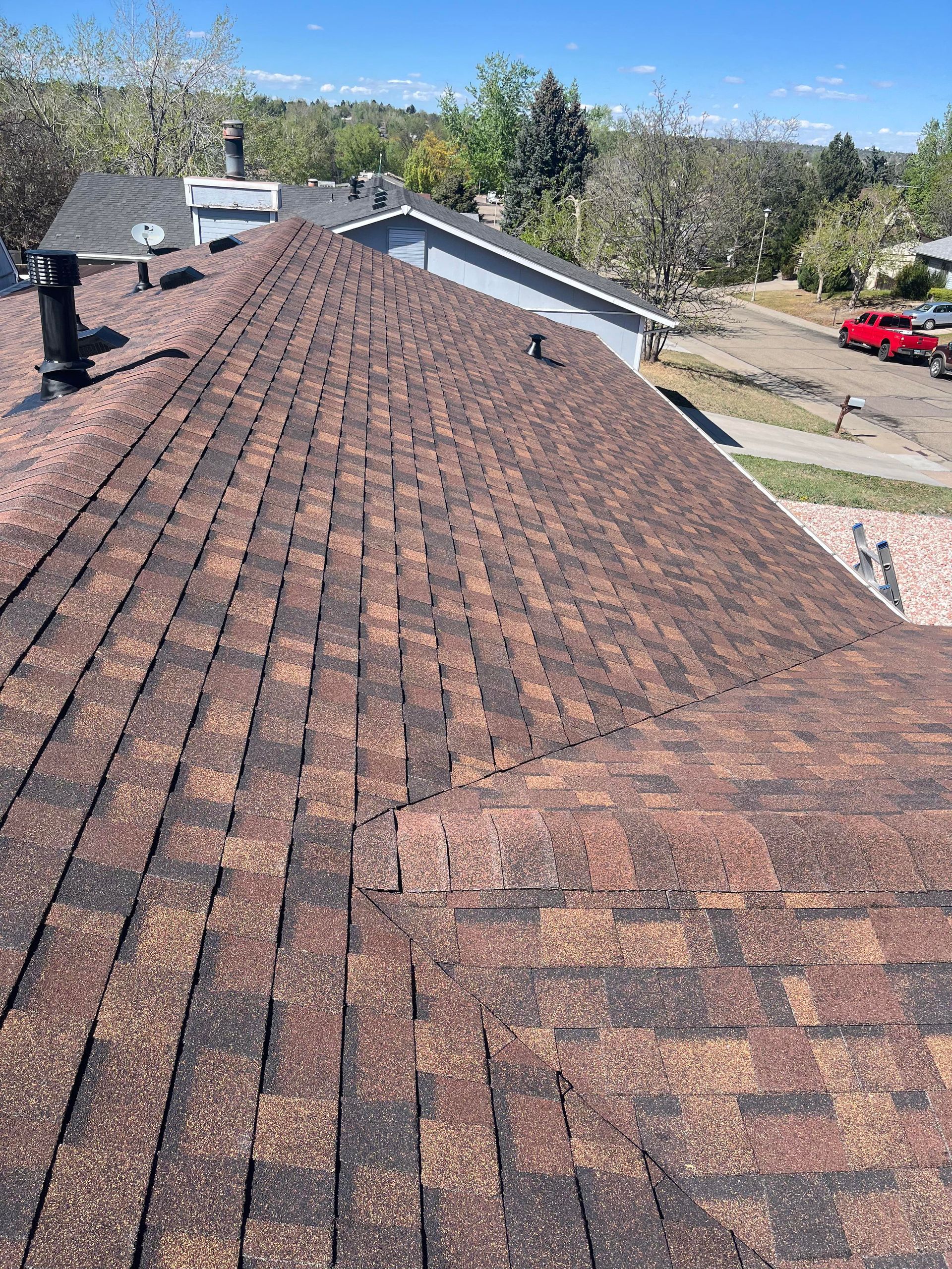 Shingled roof with a chimney, viewed from above, overlooking trees, houses, and a parked red truck.
