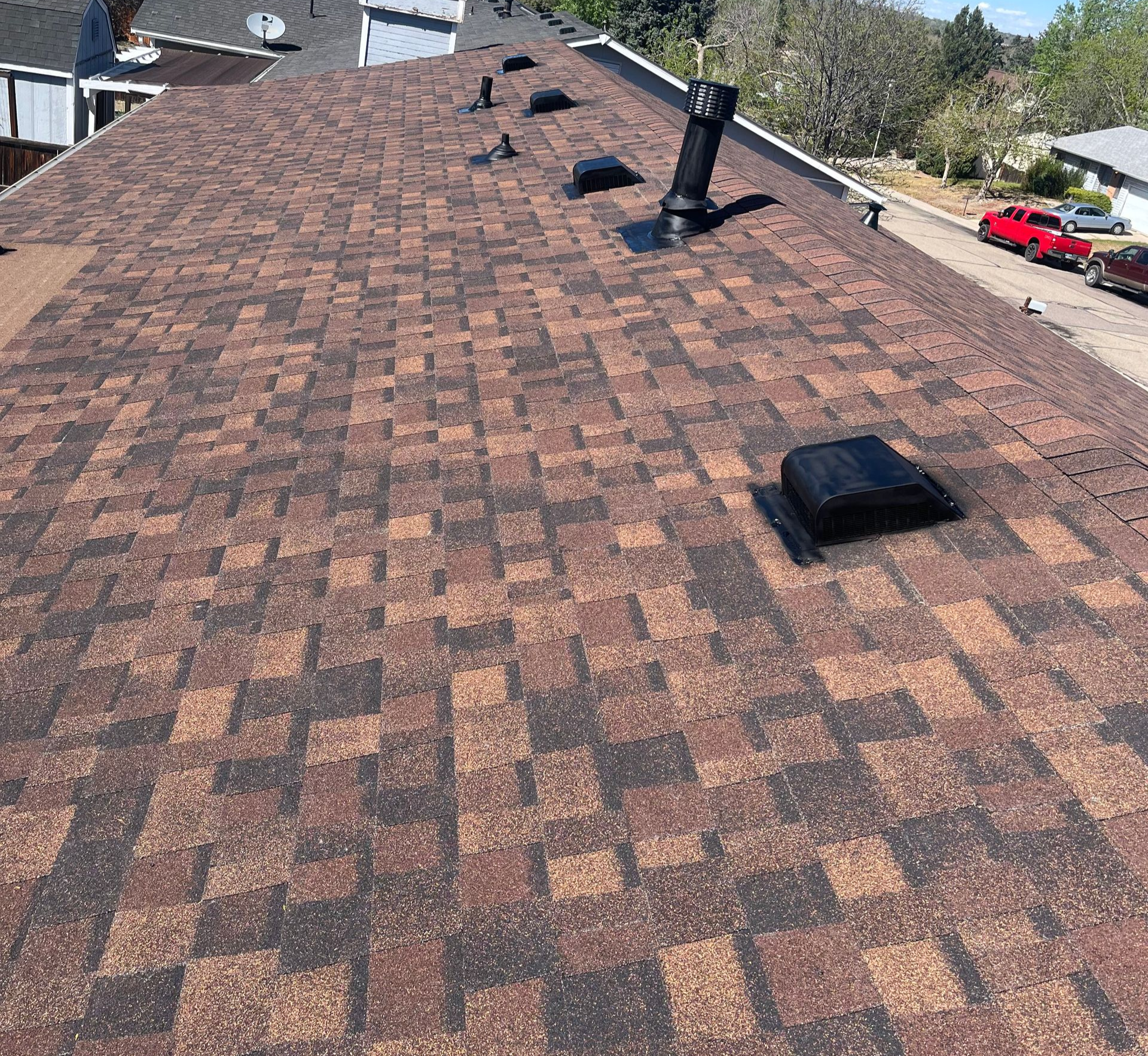 Rooftop view of brown shingle roof with vents and skylights near parked cars on a street
