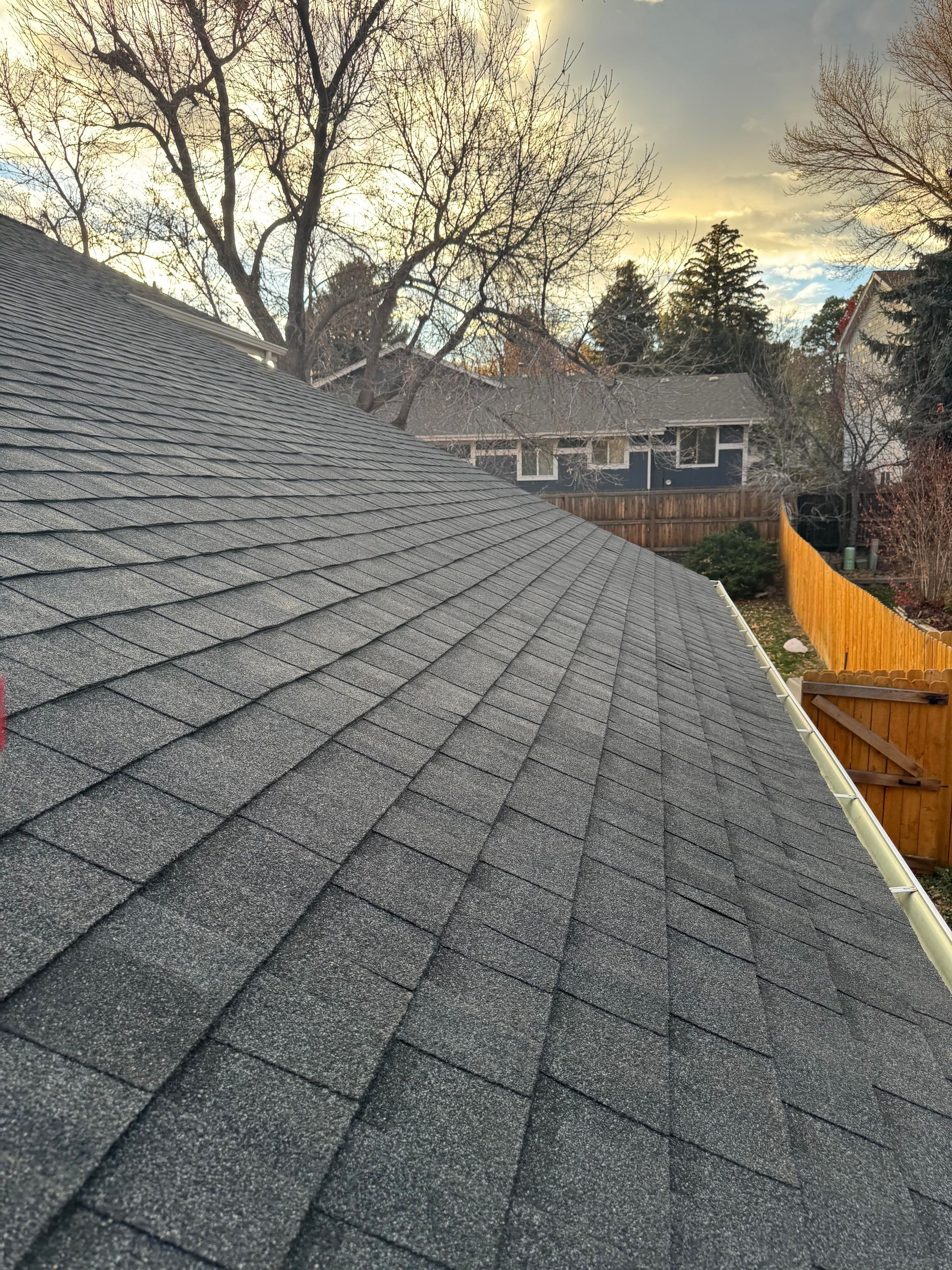 Steep gray shingled roof at sunset, with trees, neighboring house, and a wooden fence in the background