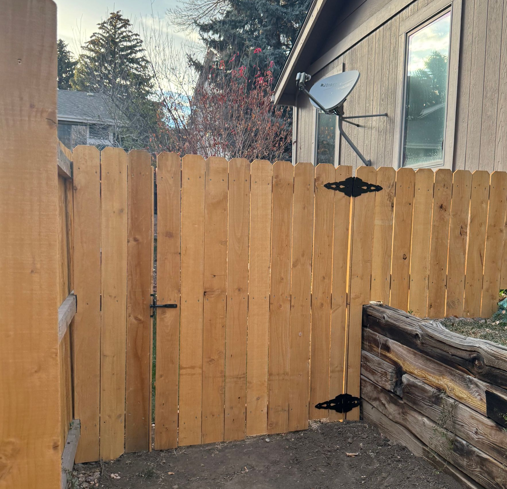 Backyard wooden fence with gate beside a house, with a satellite dish and raised planter in view