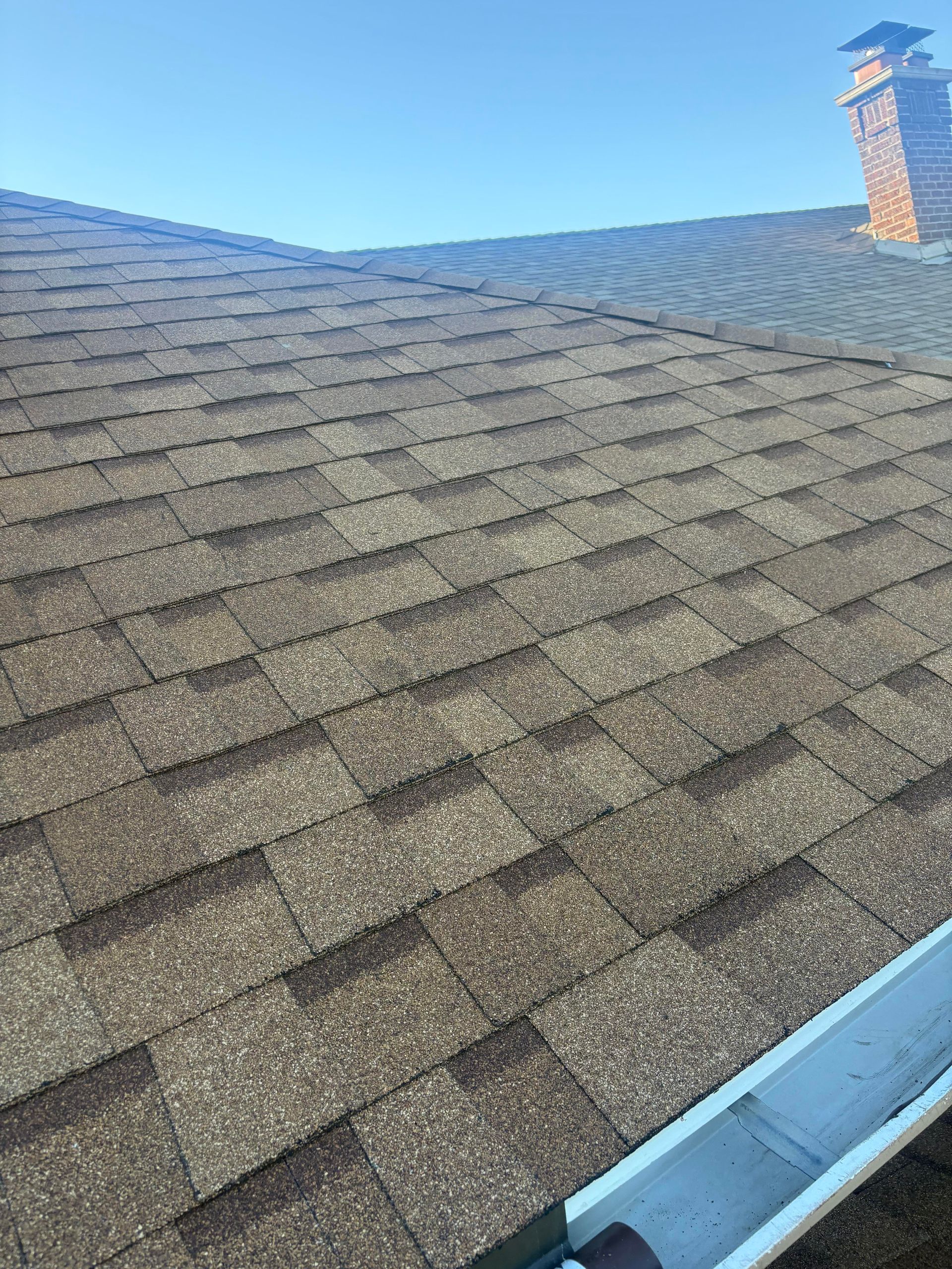 Brown shingled roof with a chimney against a blue sky, viewed from below