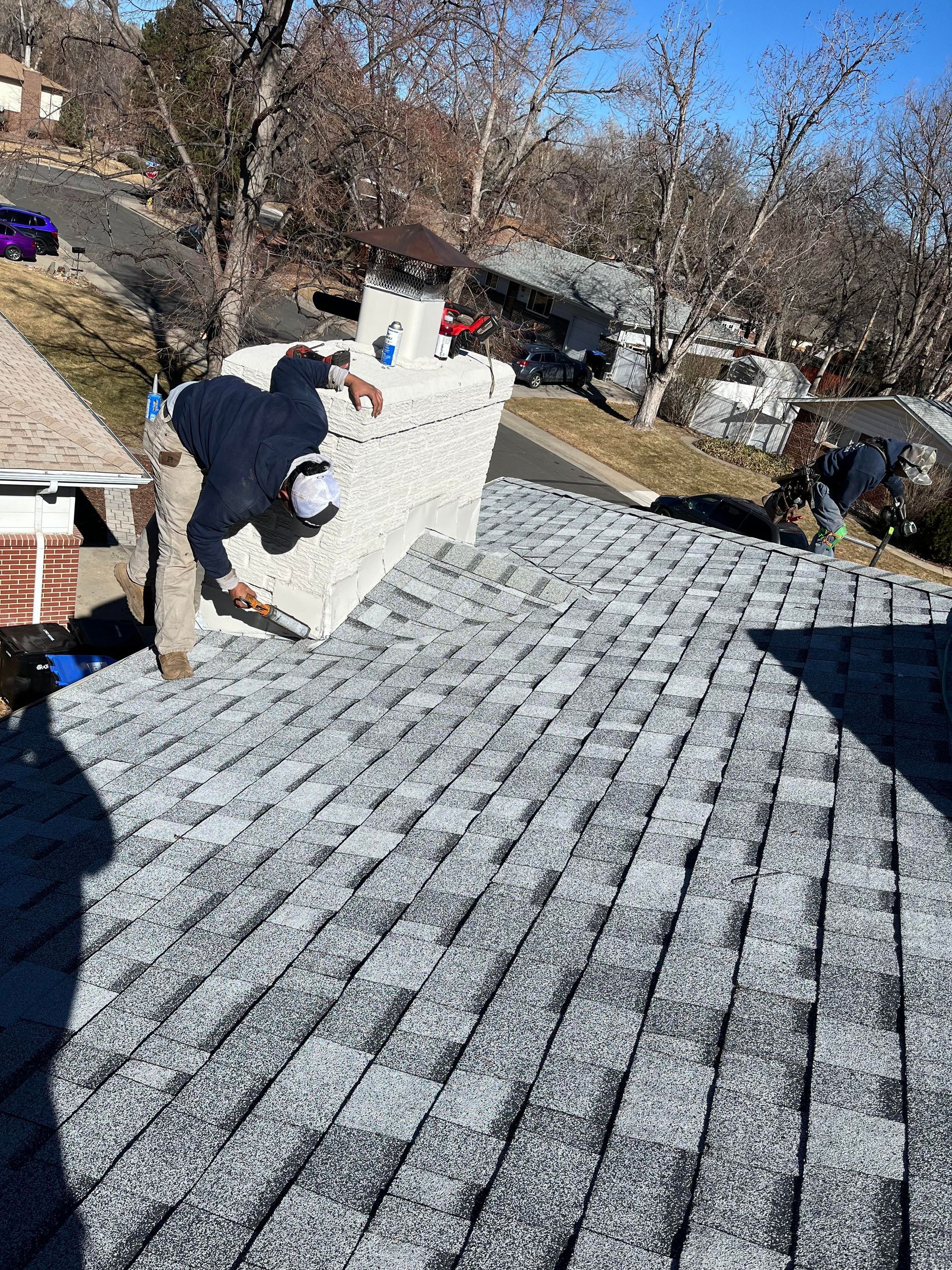 Worker on a snowy roof installing or inspecting shingles near a brick chimney.