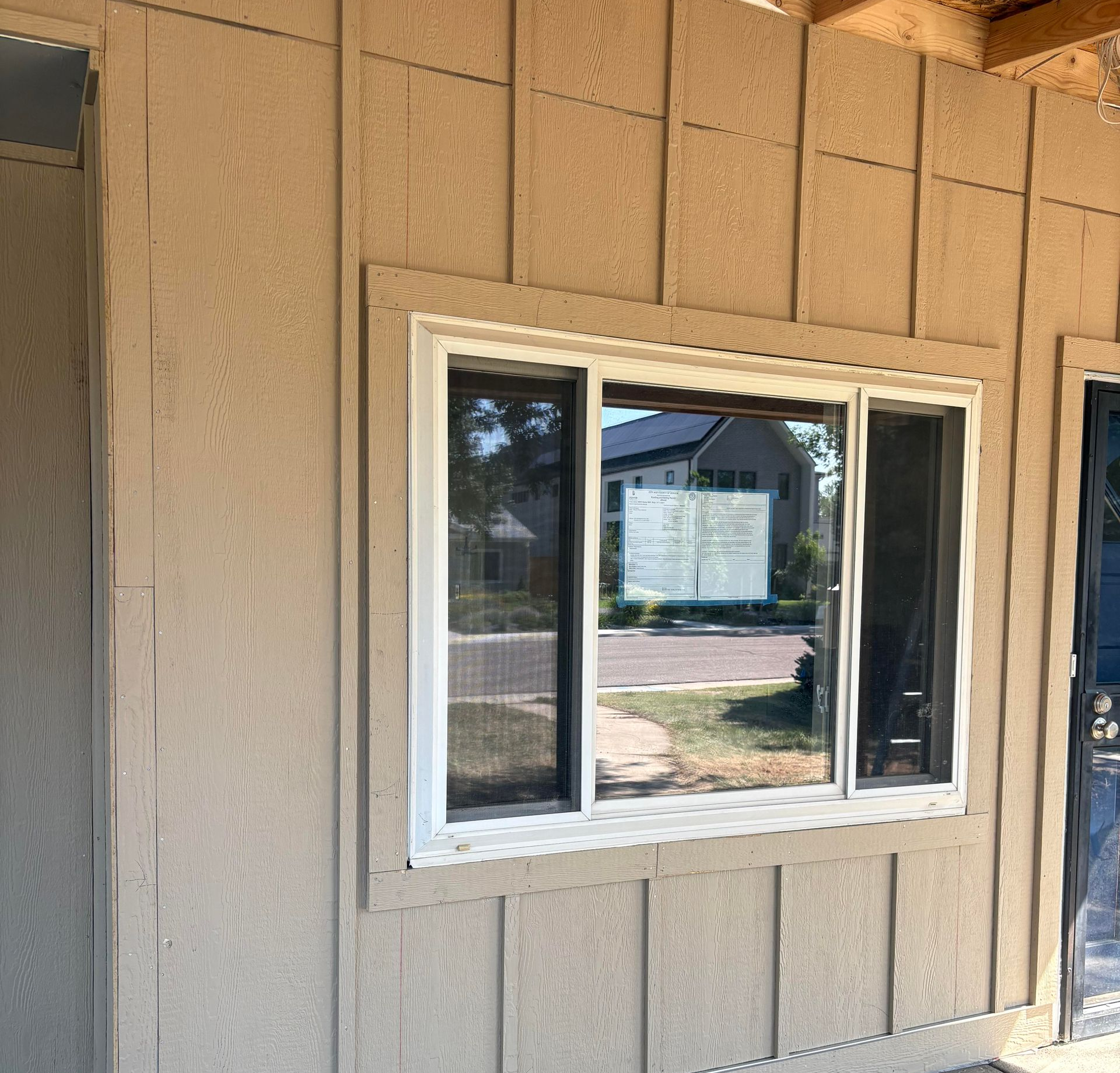 Tan siding exterior with a white-trimmed window reflecting a street and house across the road