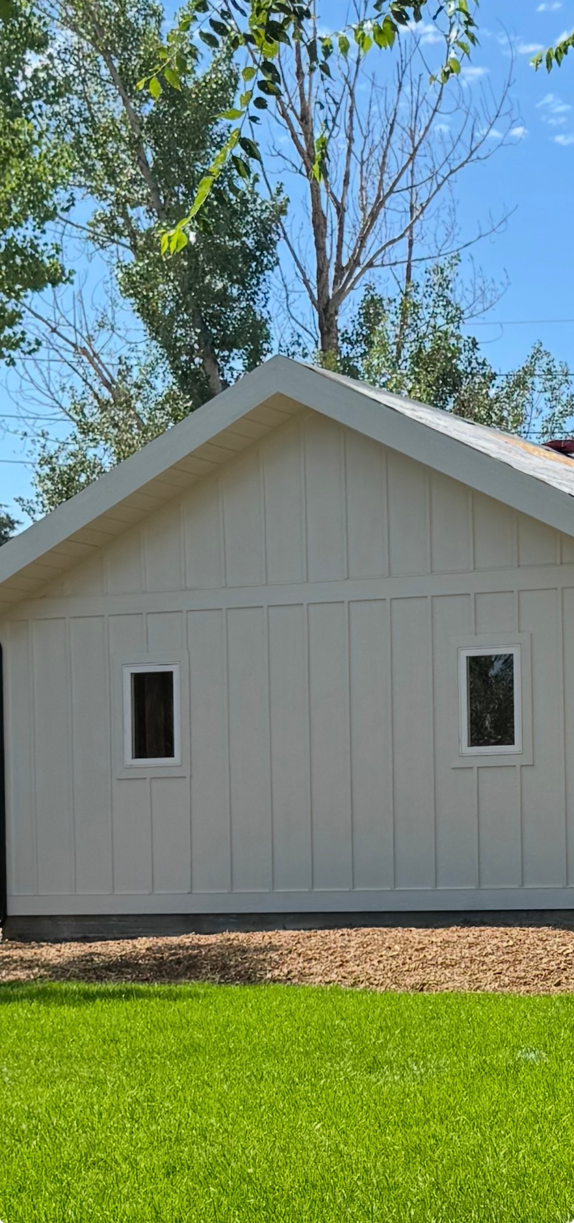 White shed with vertical siding and small windows, set on grass beneath tall trees under a blue sky