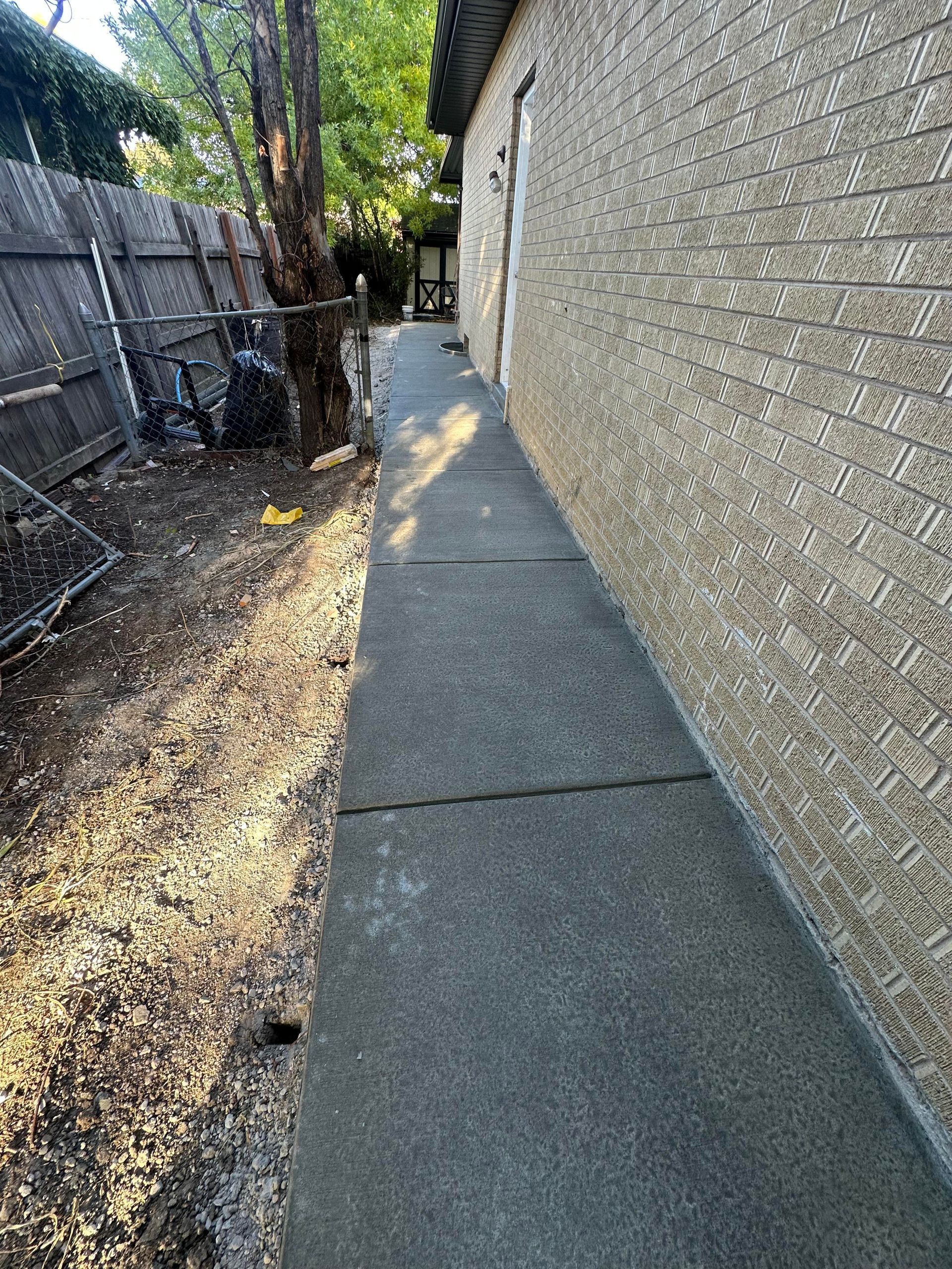 Narrow concrete side path beside a stone wall and chain-link fence, with tree shadows and dry leaves.