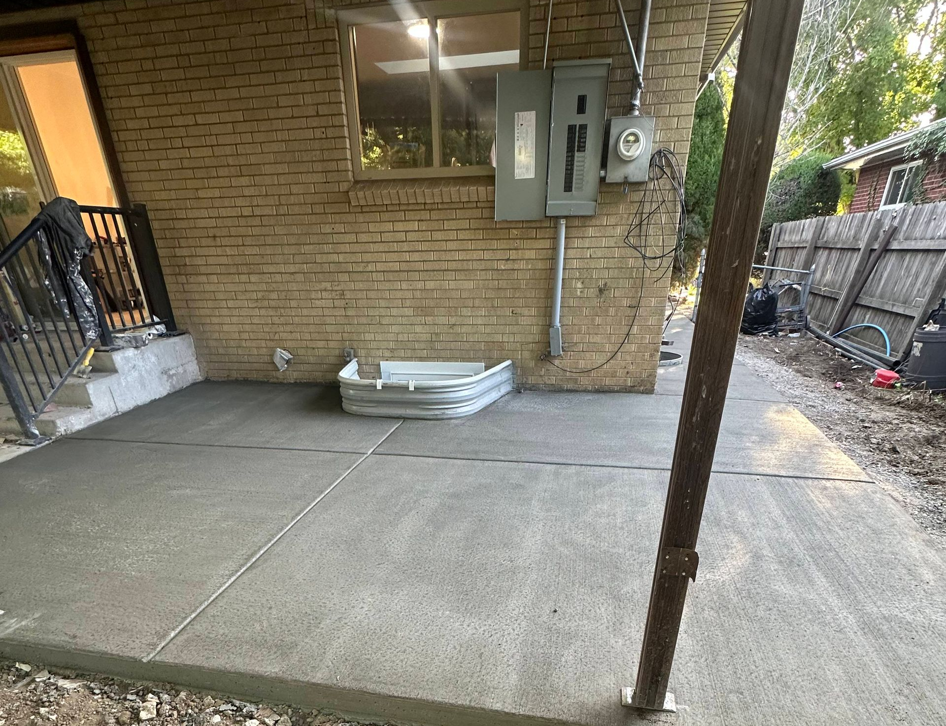 Concrete patio beside a brick house with a utility meter, stairs, and a stack of white boards.