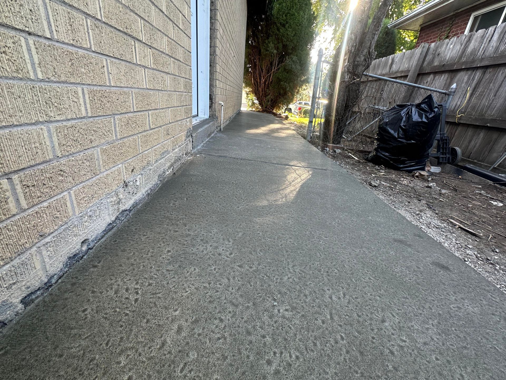 Narrow concrete walkway beside a brick wall and wooden fence, with sunlight at the far end.