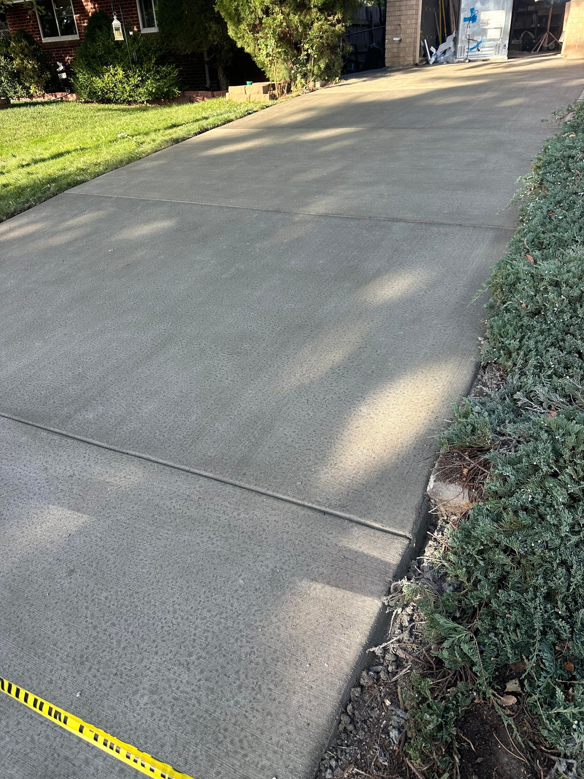 Driveway with cracked concrete, bordered by grass and shrubs, with a yellow tape measure at the bottom edge