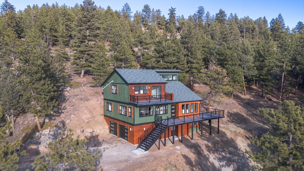 Two-story green cabin with deck on a rocky hillside, surrounded by dense pine forest