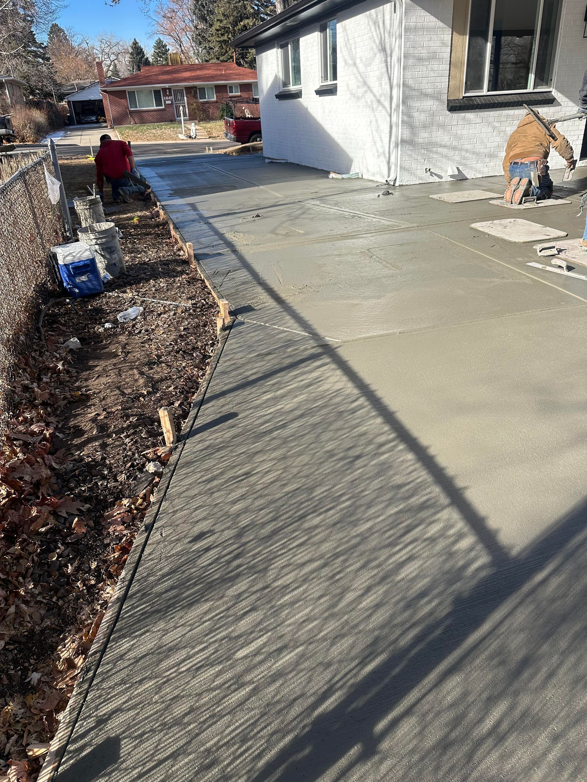 Concrete walkway beside a white house, with a person crouching near the wall and another in a red jacket in the yard.