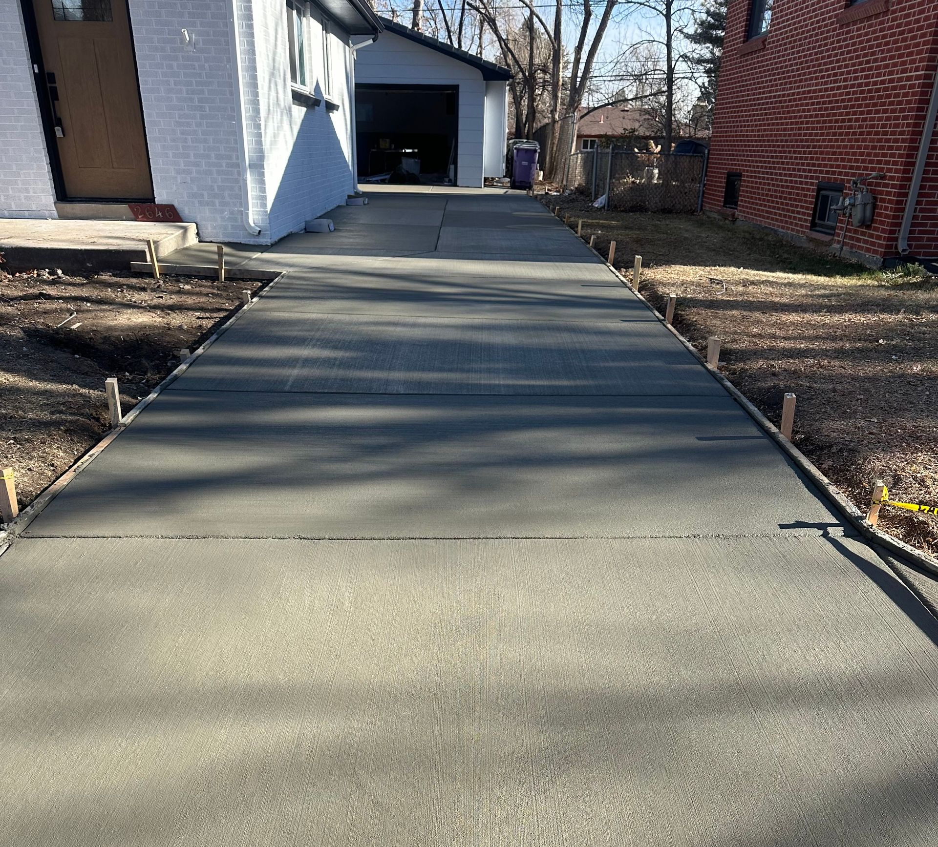 Concrete driveway leading to a garage beside a white house and brick wall, with bare trees in the background