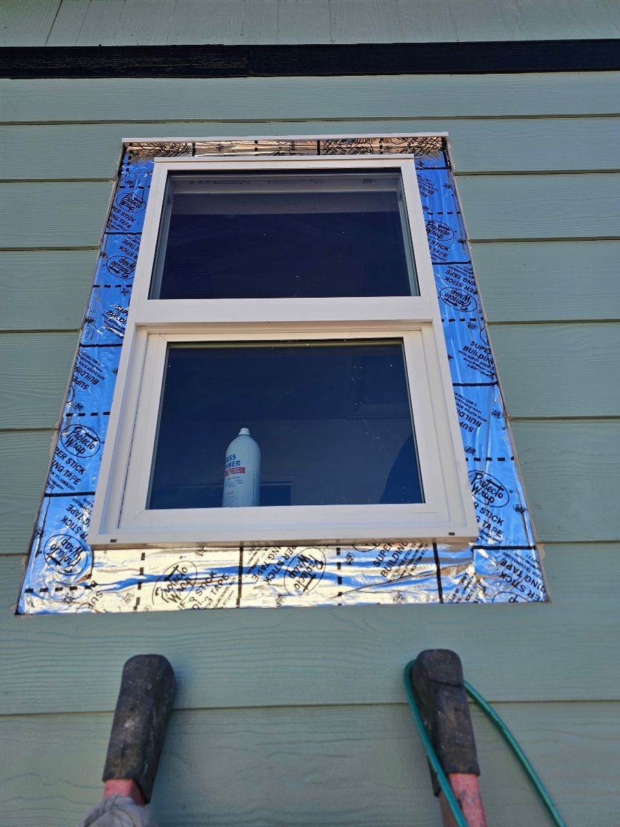 Small window with blue patterned trim on green siding, with a bottle visible inside the lower pane.