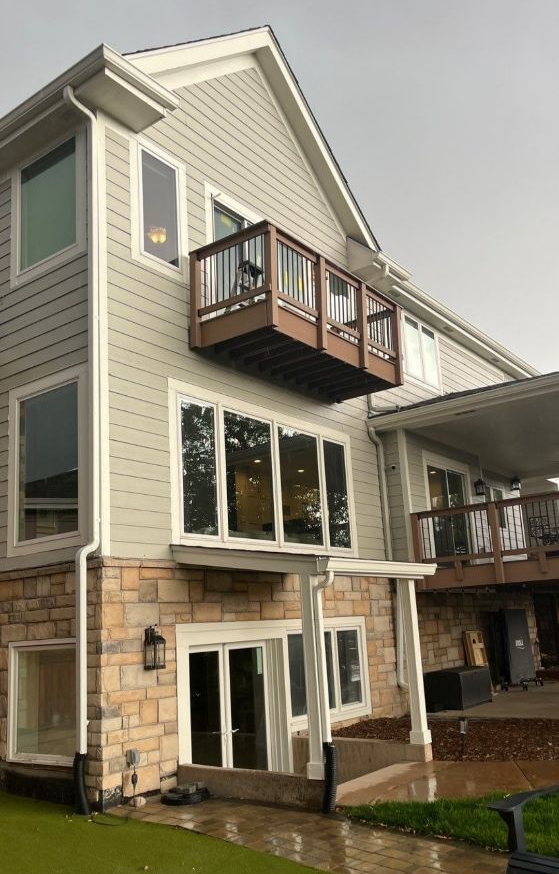 Modern two-story house with beige siding, stone base, balconies, and a covered patio during rain