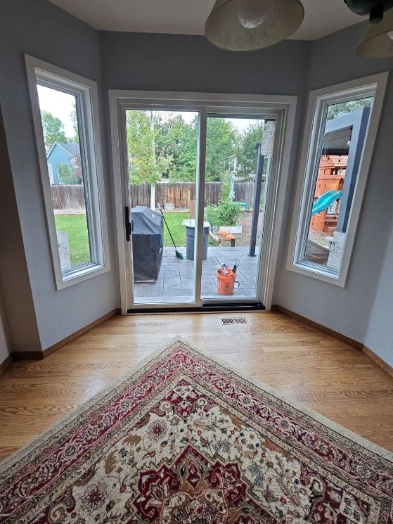 Bright sunroom with bay windows, glass doors, hardwood floor, and patterned rug overlooking a backyard.