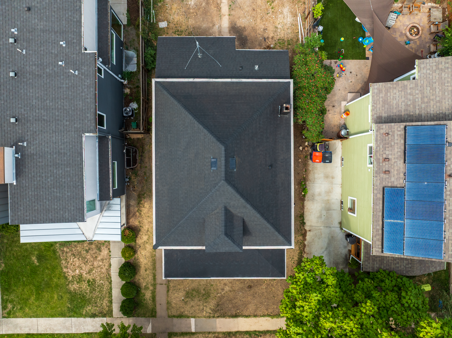 Aerial view of a dark-roofed house between neighboring homes, with a driveway and backyard.