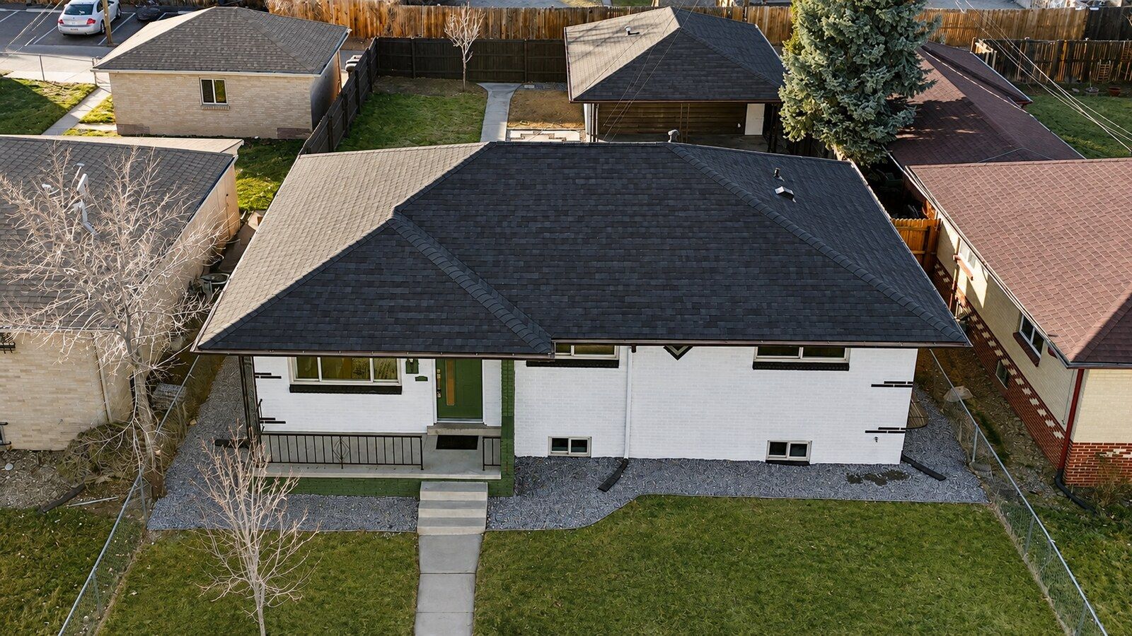 Aerial view of a small white house with a dark roof and front lawn in a suburban neighborhood.