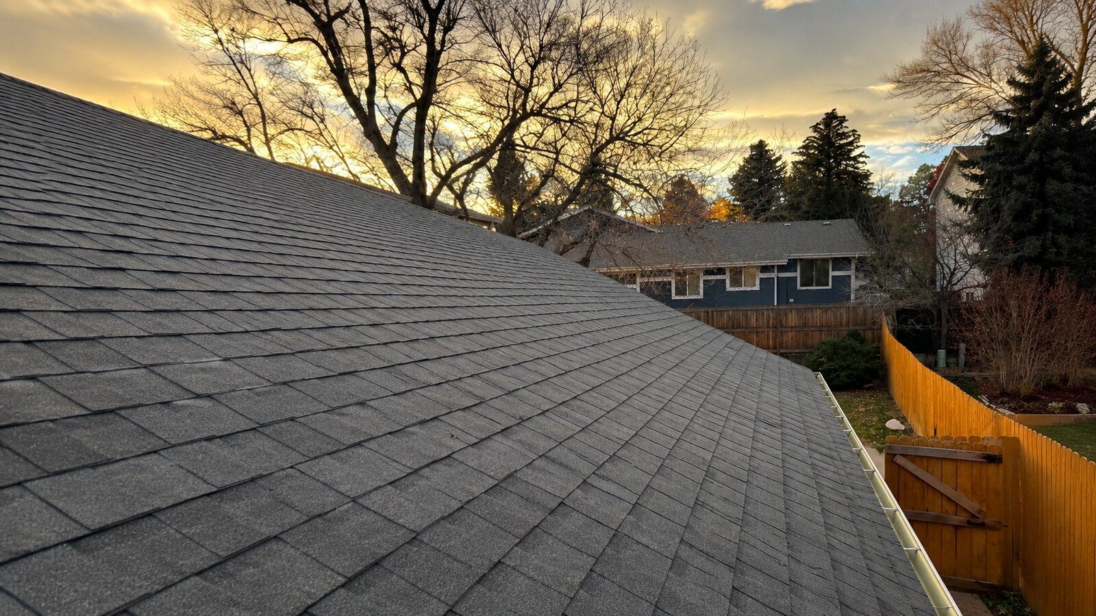 Gray shingled roof at sunset, with trees, neighboring house, and wooden fence in the background