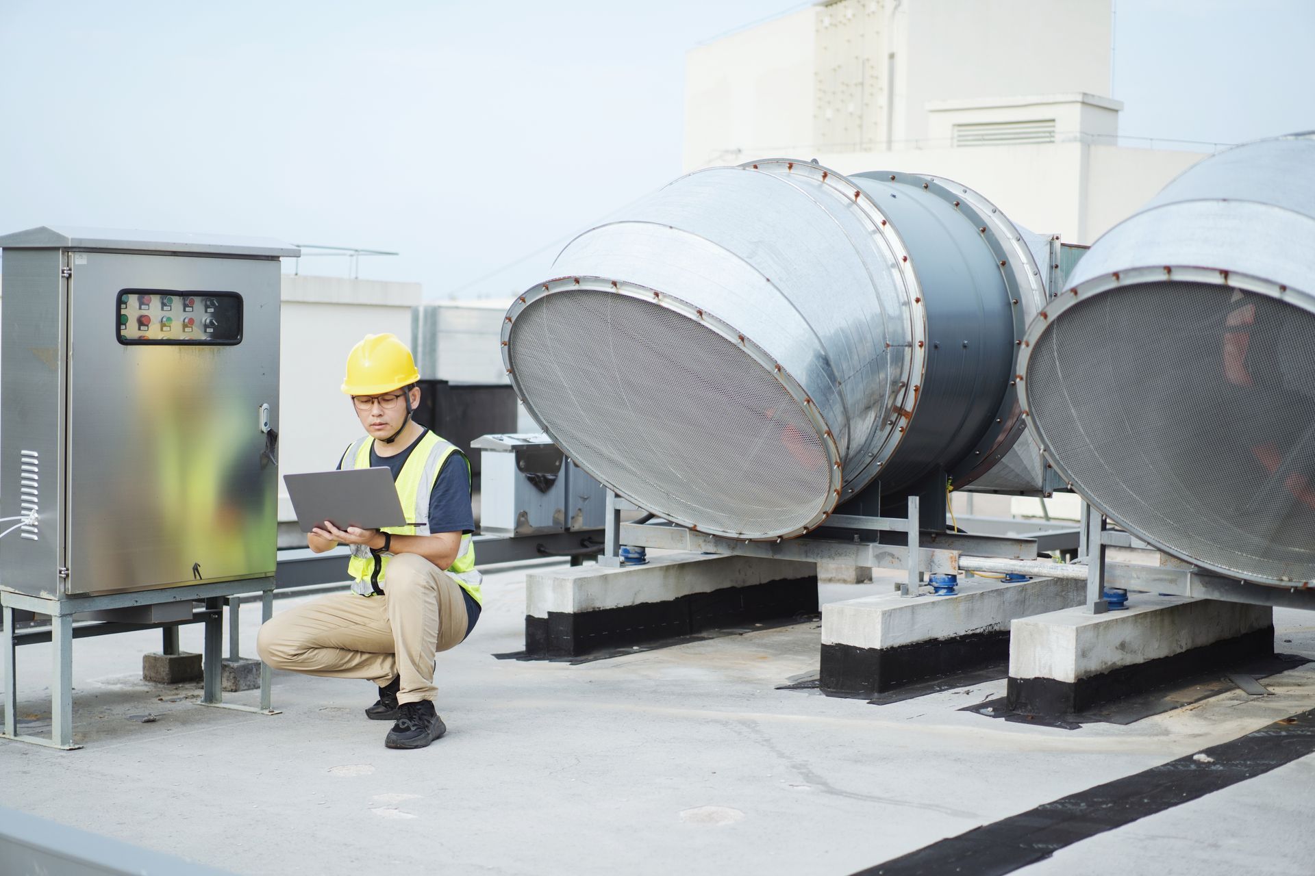 HVAC technician inspecting machinery on a rooftop with a laptop. Wearing safety vest and hard hat. Engineer in hard hat inspects HVAC system on a rooftop, using a laptop near metal ventilation equipment.