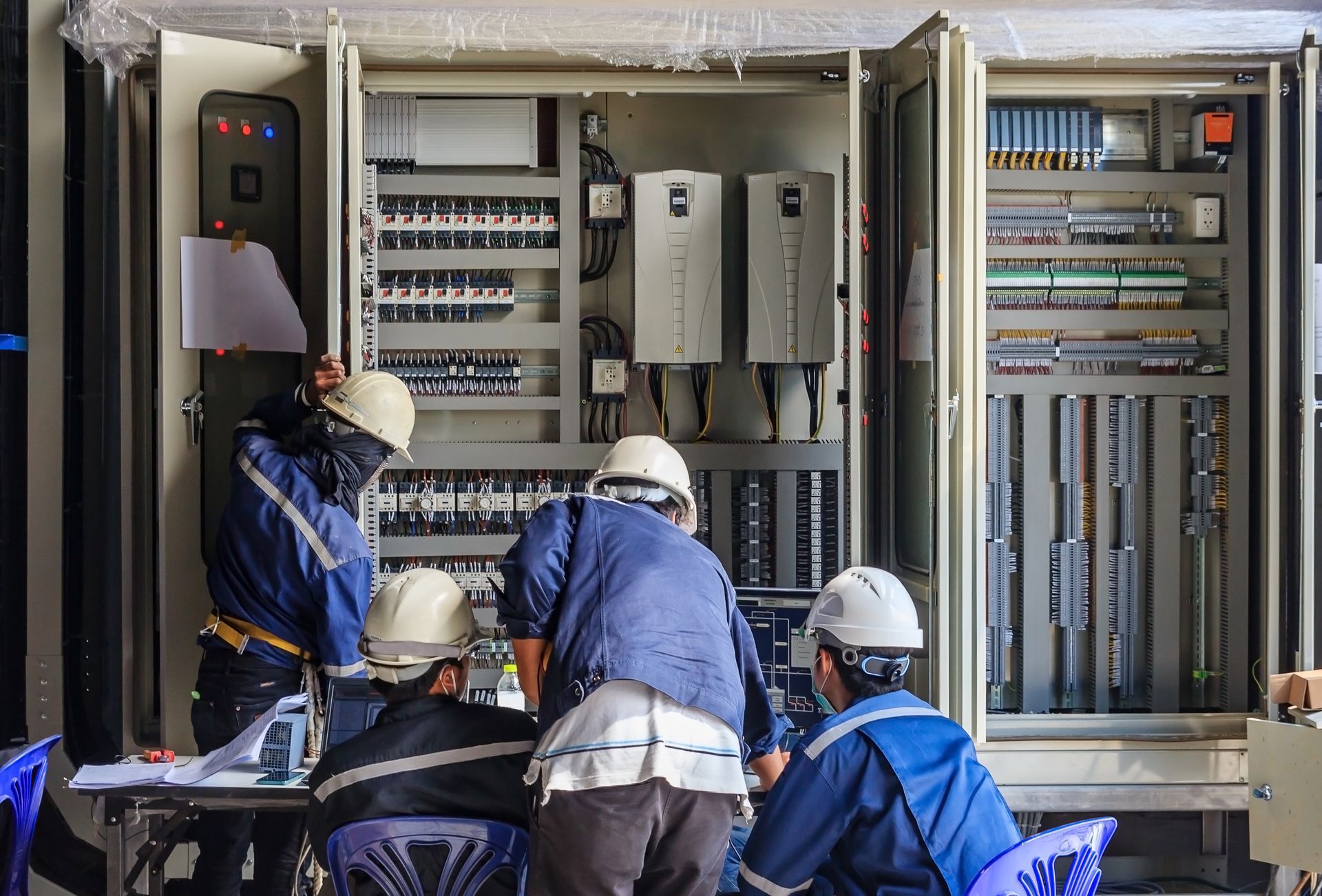 Electrical workers in blue coveralls and hard hats working on an open control panel.