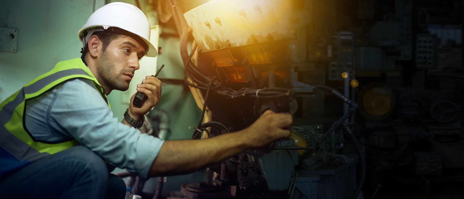 Man in work clothes and hard hat with a walkie-talkie inspects equipment in a dim industrial setting.