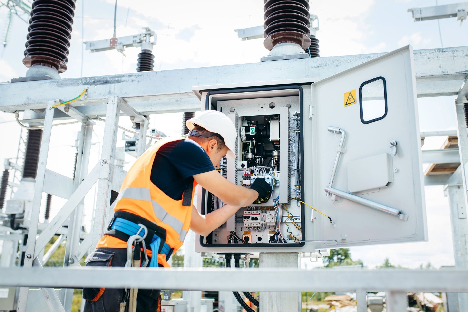 Electrician in safety gear working on a control panel at a power station. Electrician in a safety vest and hardhat working on electrical panel outdoors.