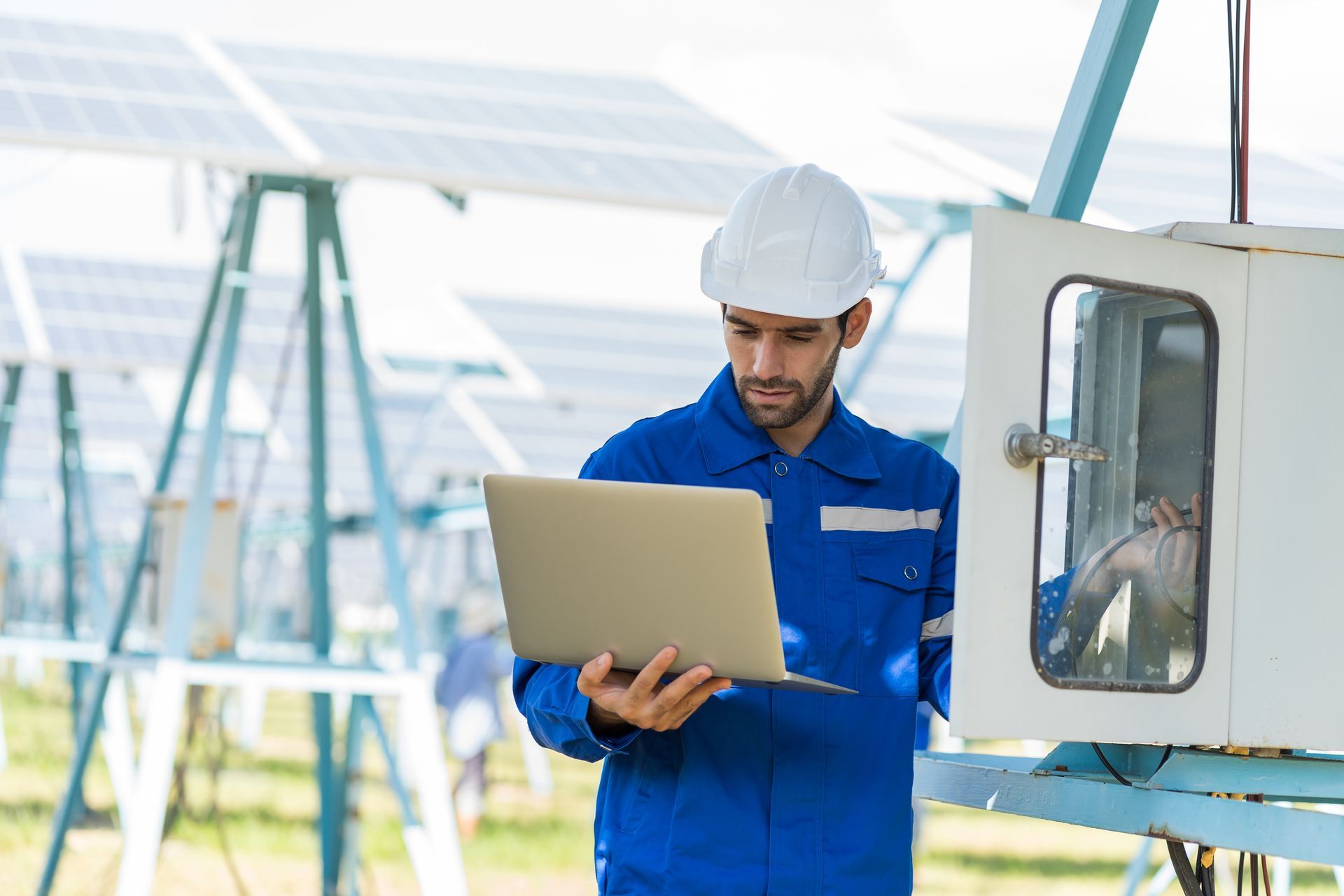 Man in blue jumpsuit and hard hat using a laptop to inspect electrical equipment at a solar panel farm. Technician in blue coveralls and hard hat uses laptop near solar panels and control box.