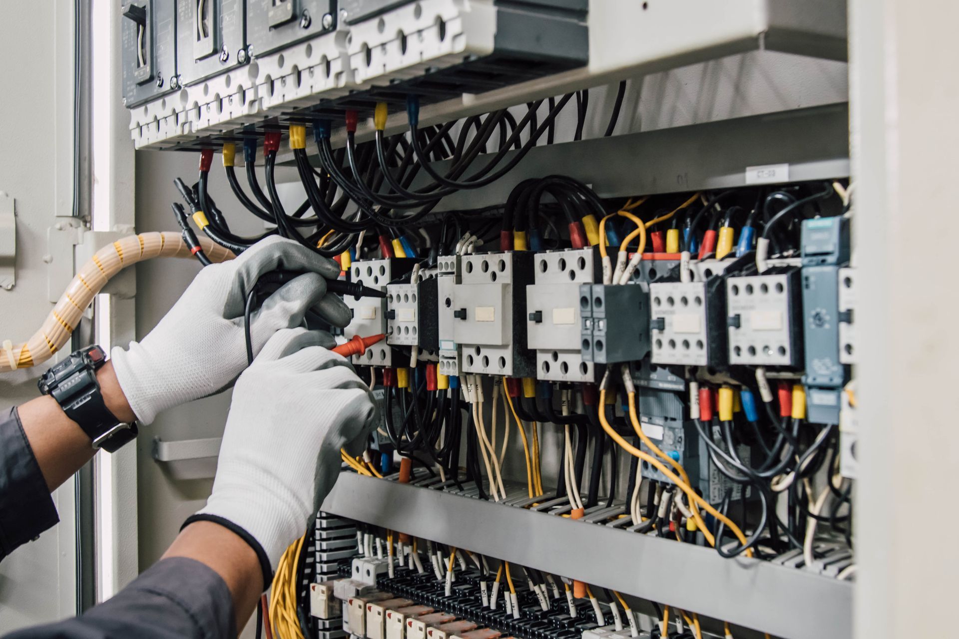 Hands wearing gloves testing electrical panel wiring. Electrician wearing gloves testing wires in a control panel with various colored cables.