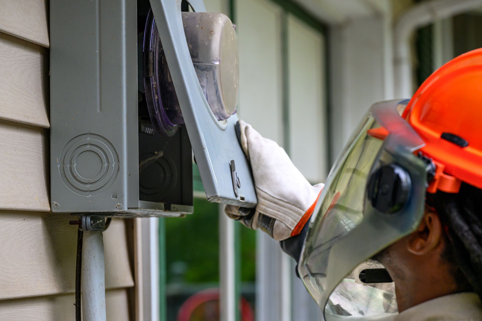 Electrician in safety gear working on a gray electrical box outside a building. Electrician in safety gear working on a gray electrical box on a beige house exterior.