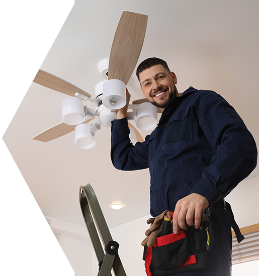 Man in blue jumpsuit installing a ceiling fan, standing on a ladder and smiling. Electrician installing a ceiling fan, standing on a ladder. He is smiling and wearing a blue uniform.