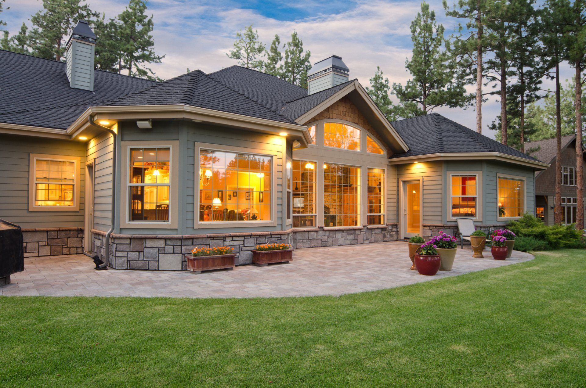Exterior view of a house with a stone patio, green lawn, and trees under a cloudy blue sky. Exterior view of a house with a stone patio, green lawn, and trees under a cloudy blue sky.