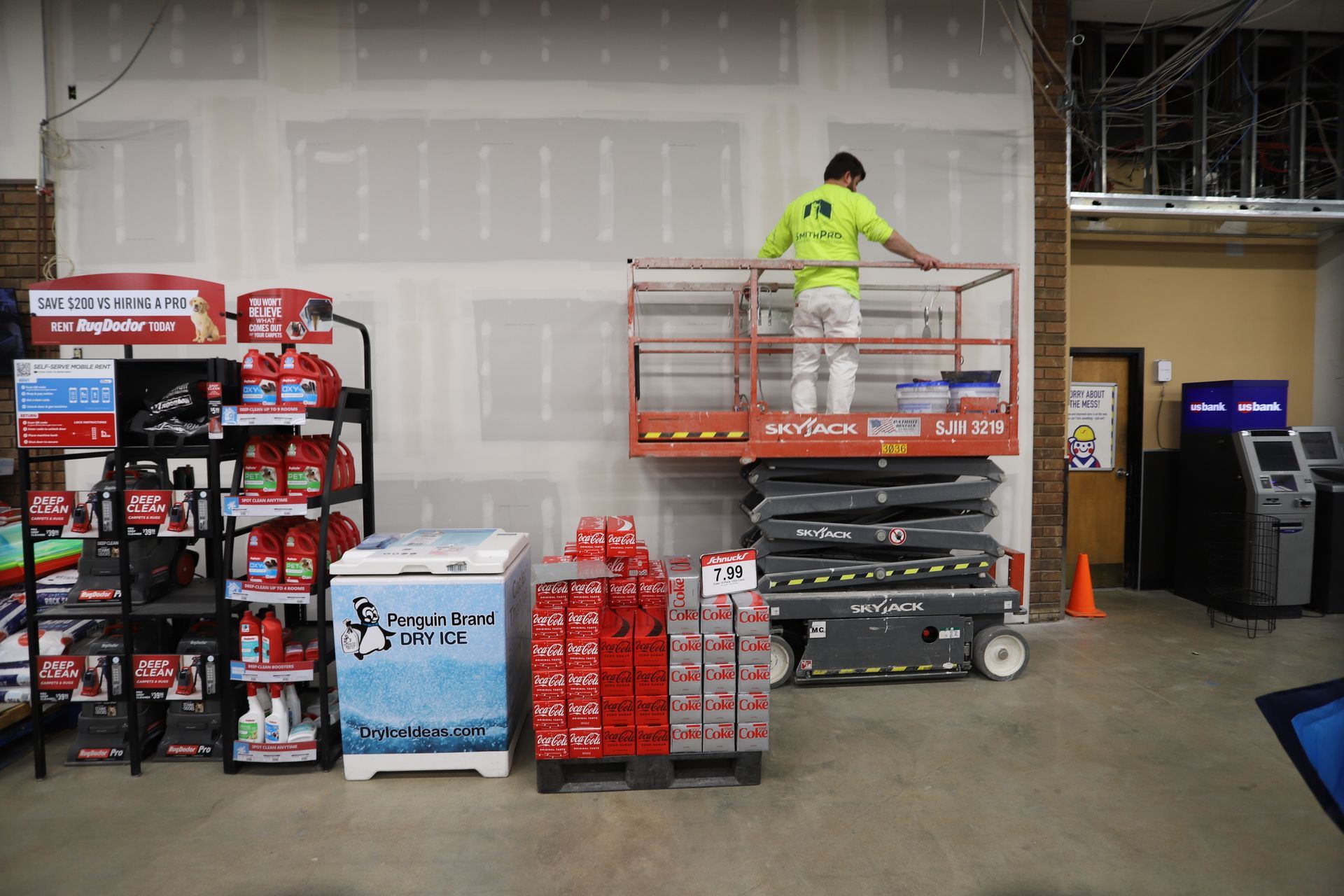Worker in neon shirt on a scissor lift beside stacked boxes in a warehouse aisle