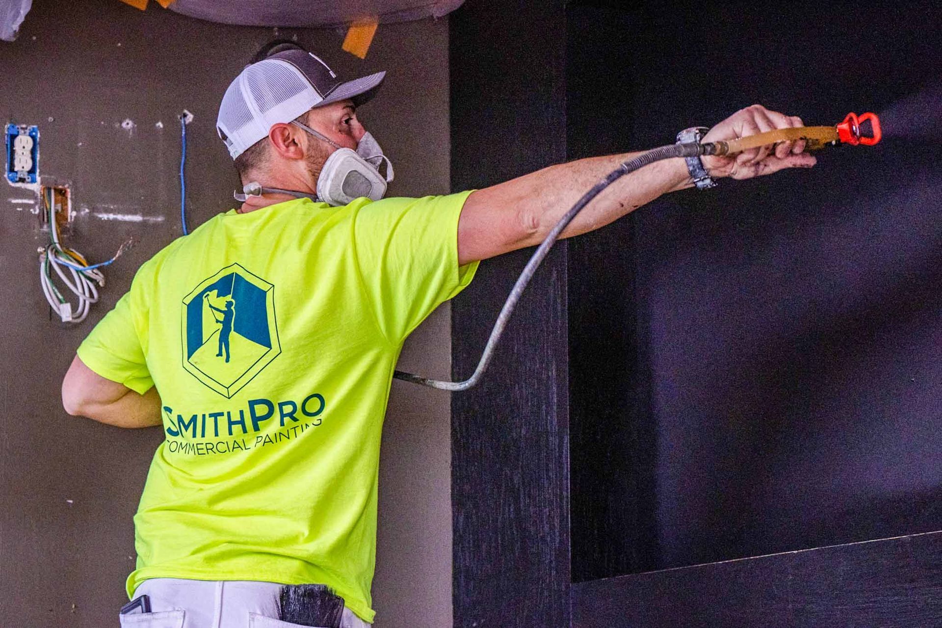 A construction worker in a neon yellow shirt and respirator mask uses a paint sprayer on a dark wall.
